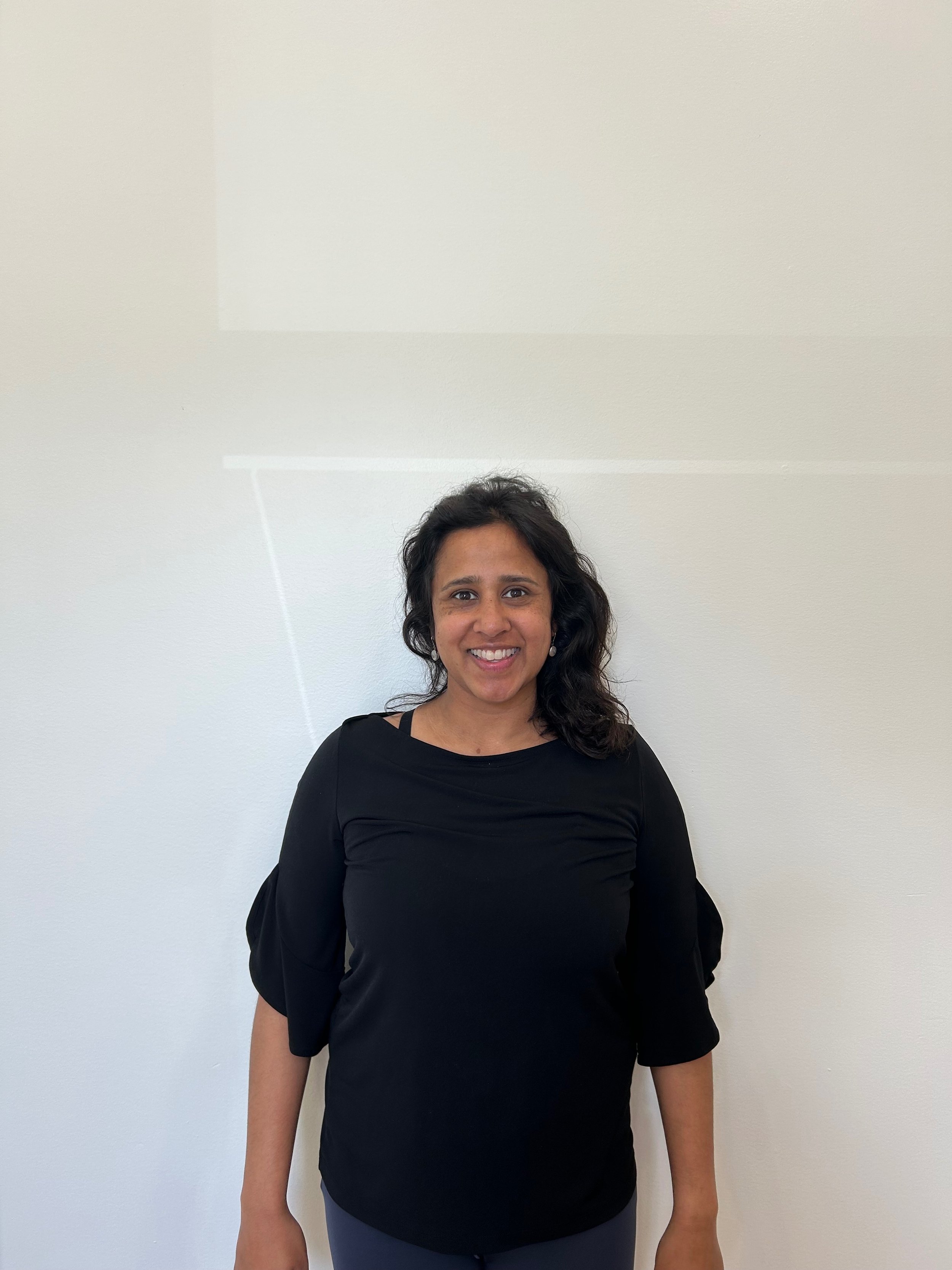 A woman with long curly black hair, smiling, wearing a black long-sleeve top and gray pants, standing in front of a plain white wall.