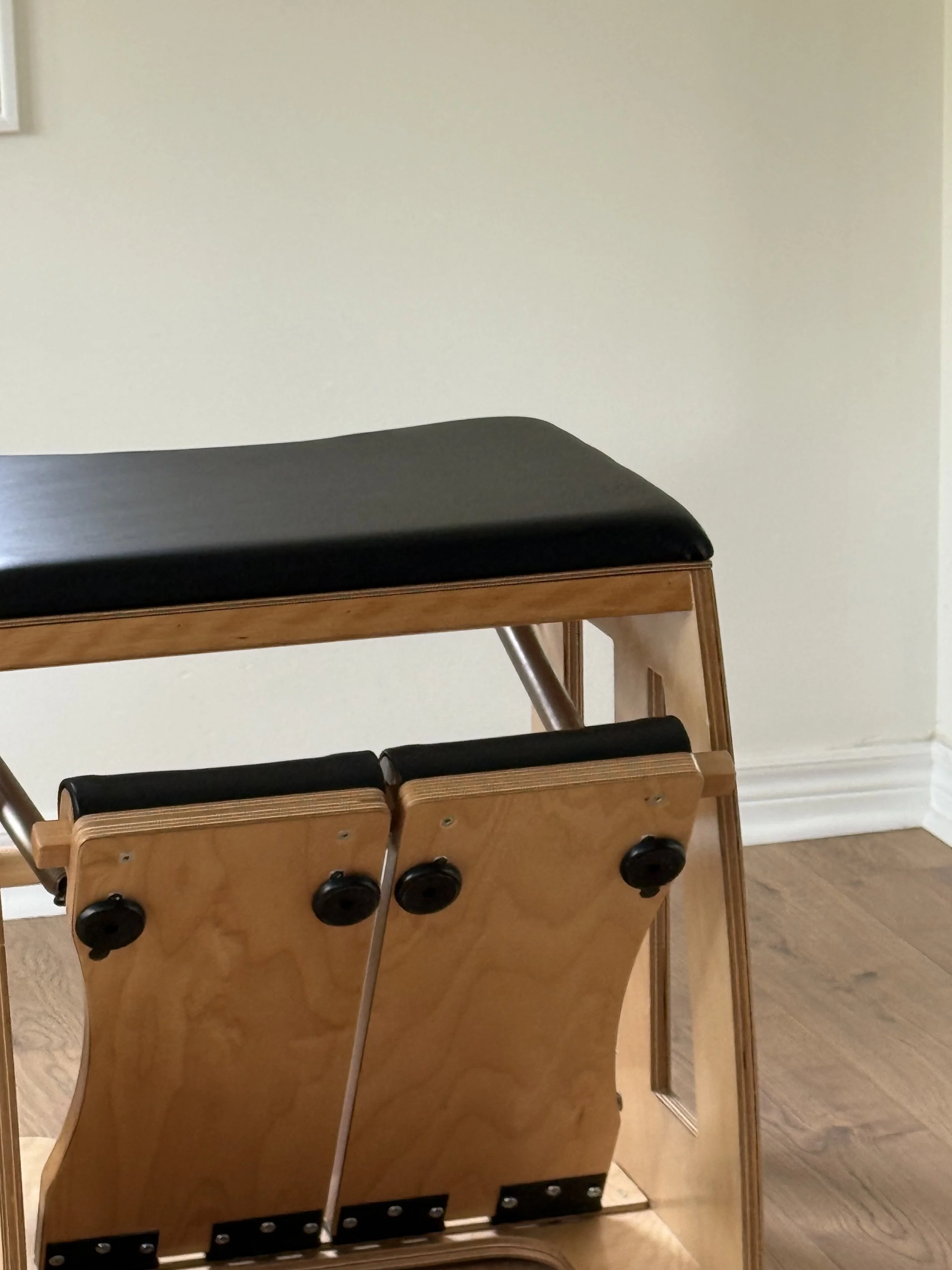 A wooden adjustable hospital or examination bed with black padded surface and controls on the side, situated against a plain off-white wall and hardwood floor.