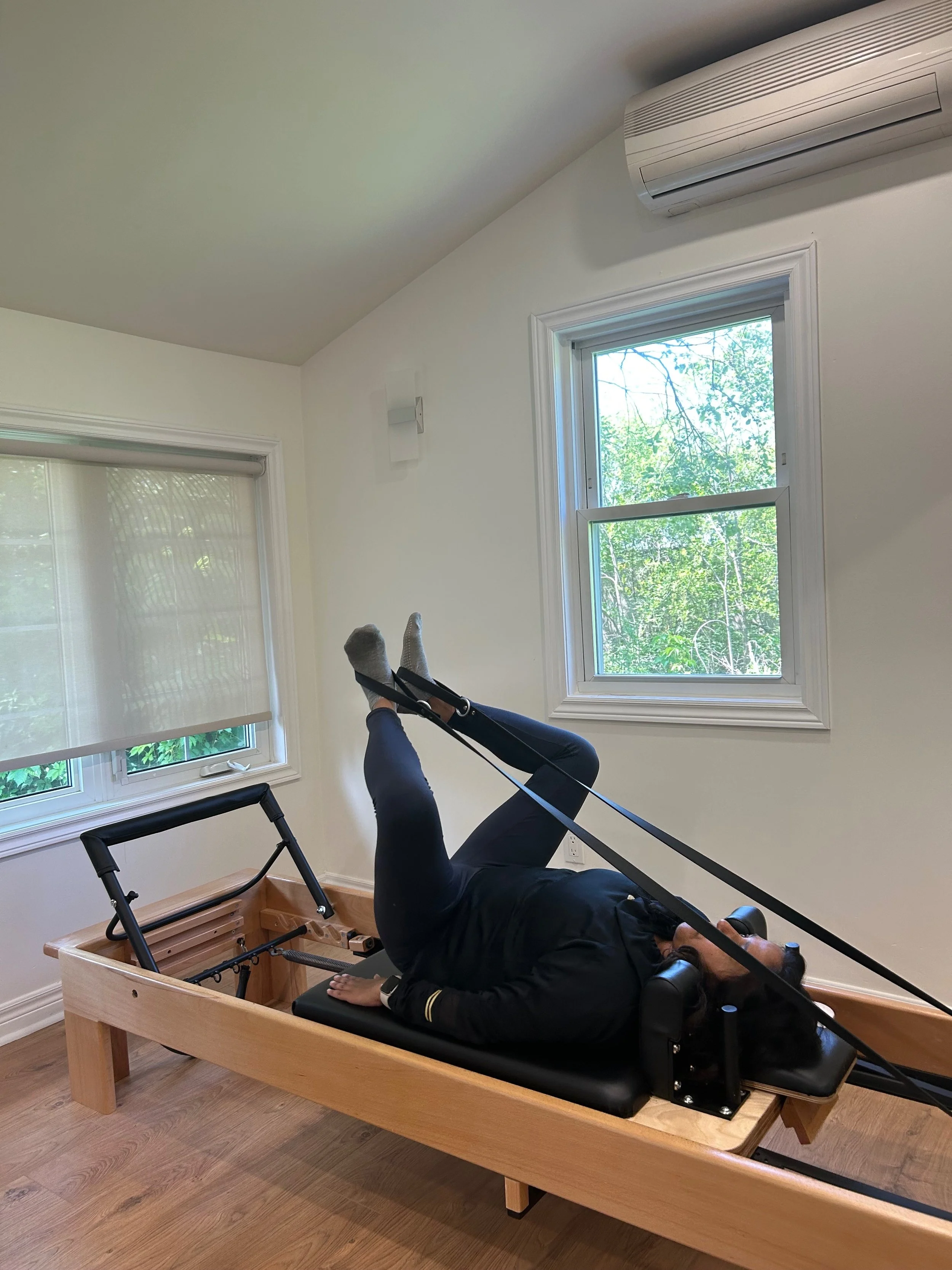 Person lying on back in a wooden Pilates reformer machine, legs raised, inside a room with two windows and an air conditioning unit.