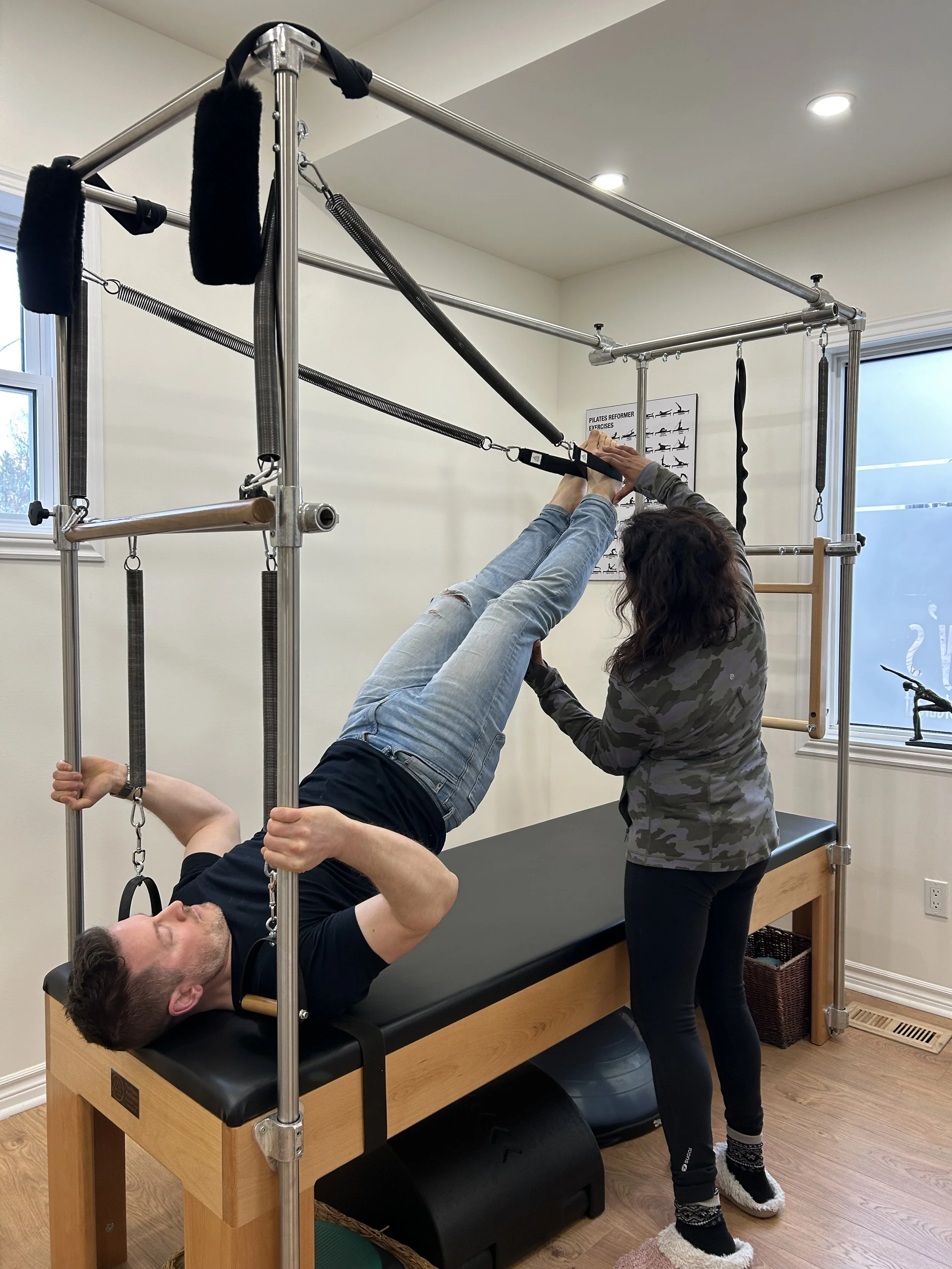 A person is lying on a massage table while a woman assists with exercises using a Pilates reformer machine. The person is gripping straps attached to the machine, and the woman is helping them with the exercise. The setting appears to be a physical therapy or Pilates studio.