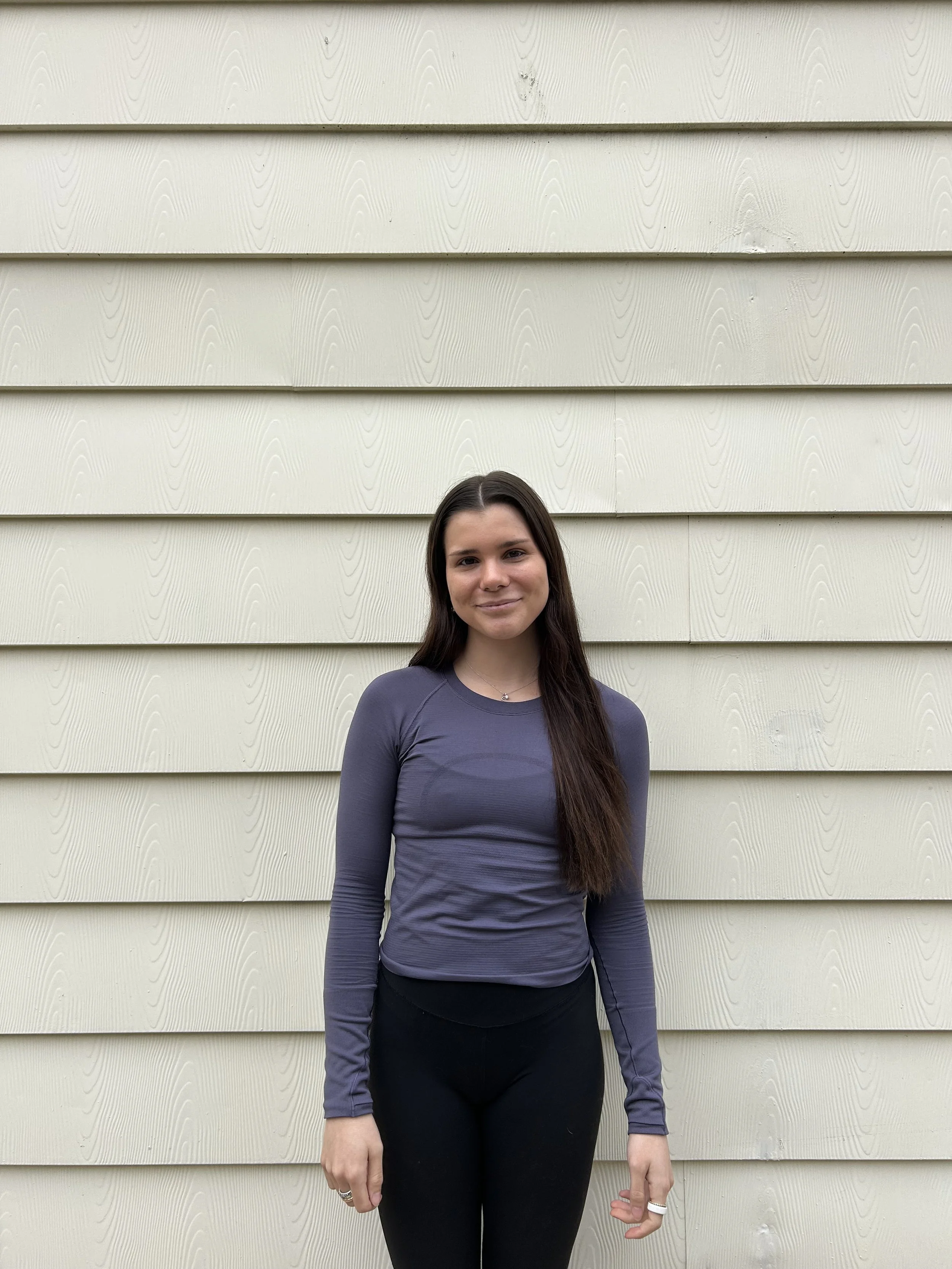 A young woman with long dark hair, wearing a dark gray long-sleeve top and black pants, standing in front of a beige, textured horizontal siding wall.