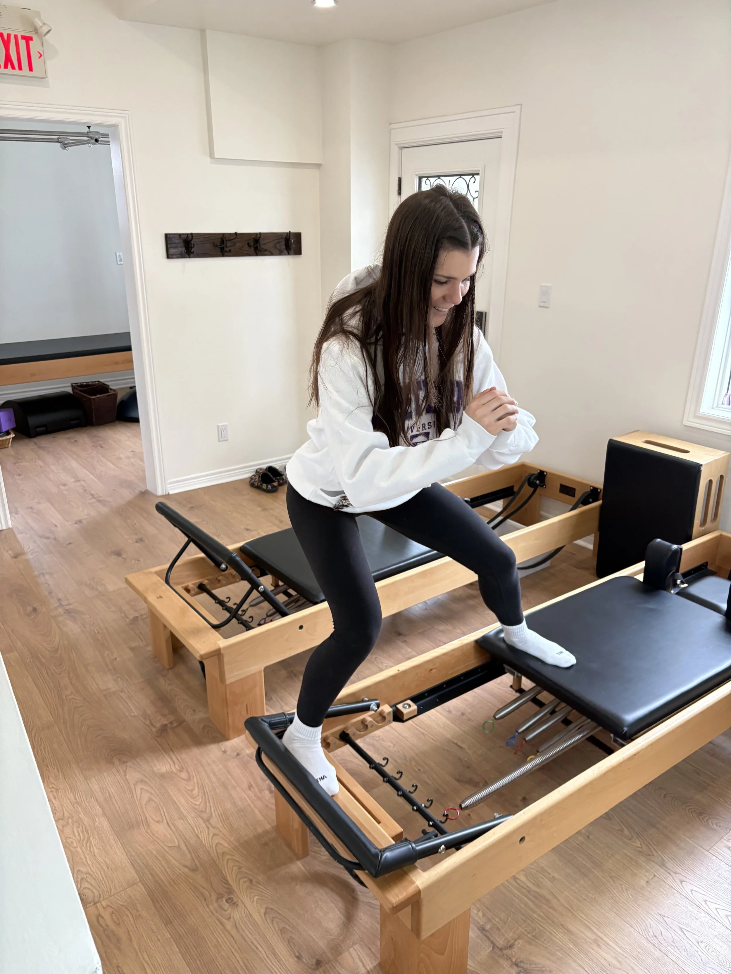 A woman with long brown hair is smiling and playing a game on a wooden activity table in a bright room. She is wearing a white hoodie, black leggings, and white socks. The room has wooden floors, white walls, a door with a small window, and a coat rack on the wall. There is an exit sign above the door.