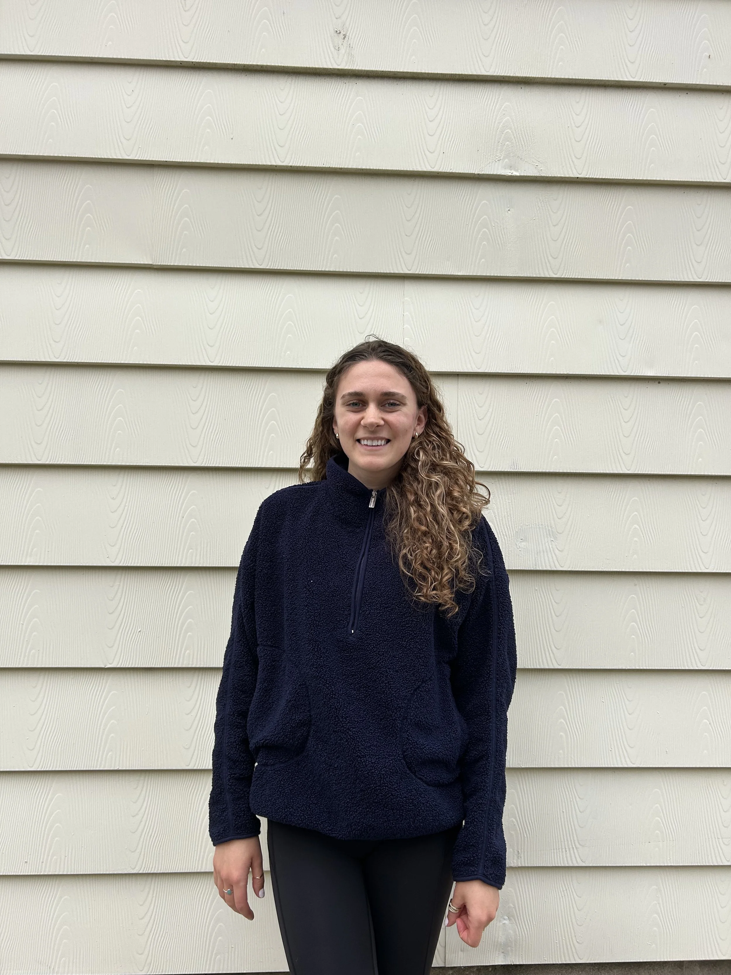 A woman with long curly hair wearing a navy blue fleece jacket and black leggings standing in front of beige horizontal siding.