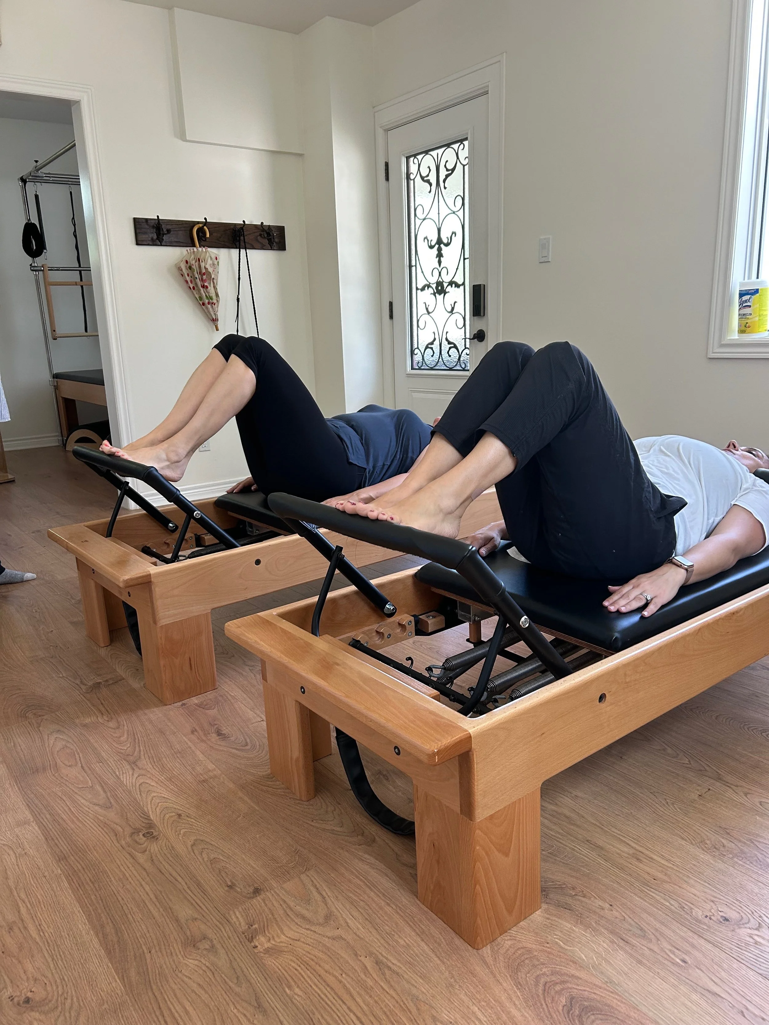 Two people are lying on chiropractic adjustable tables in a room, performing spinal adjustments, with a doorway to a walk-in closet in the background.