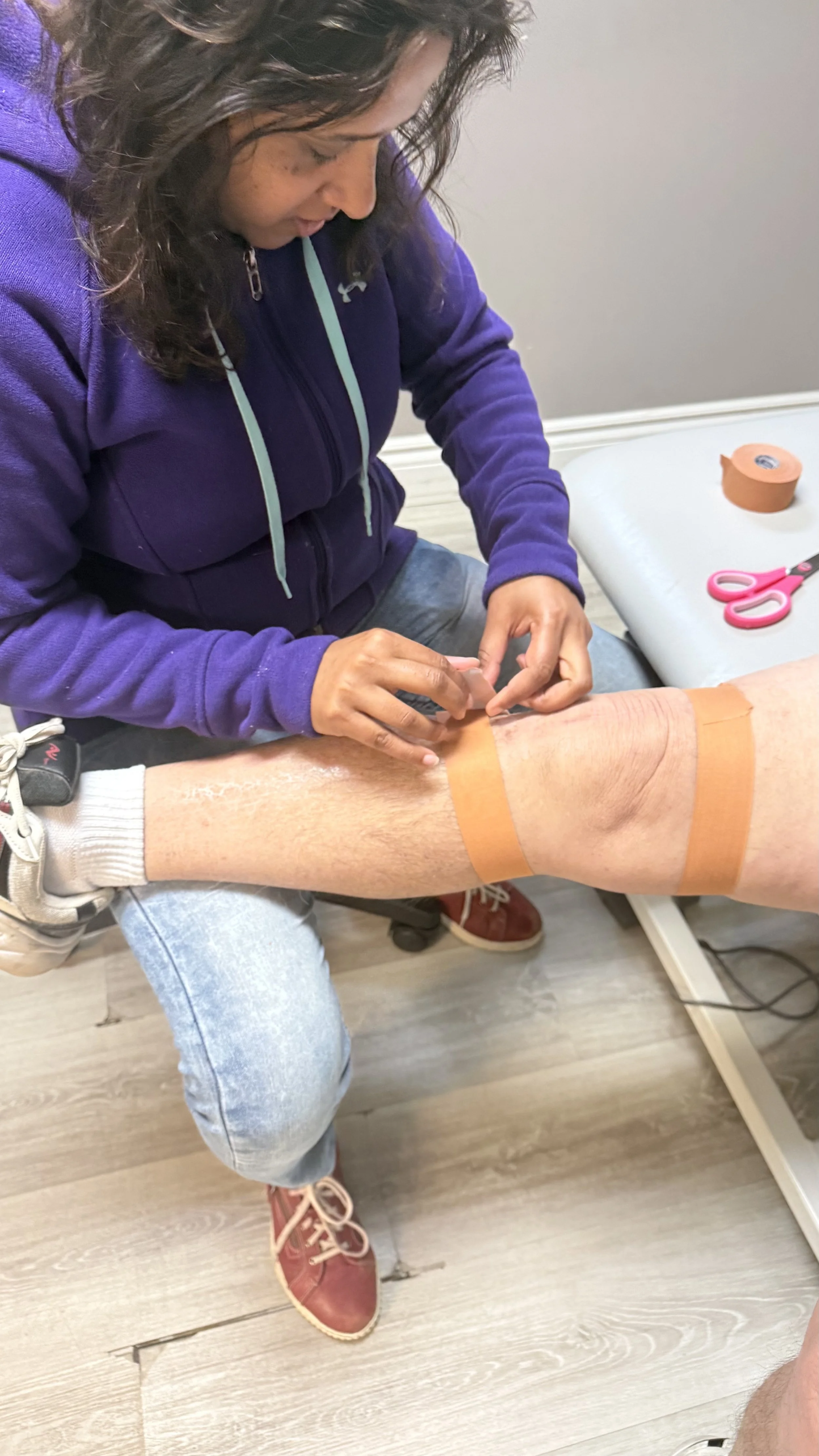 A woman in a purple jacket is applying a bandage to a person's lower leg, which is secured with orange athletic tape. There are pink scissors and a roll of tape nearby on a treatment table.