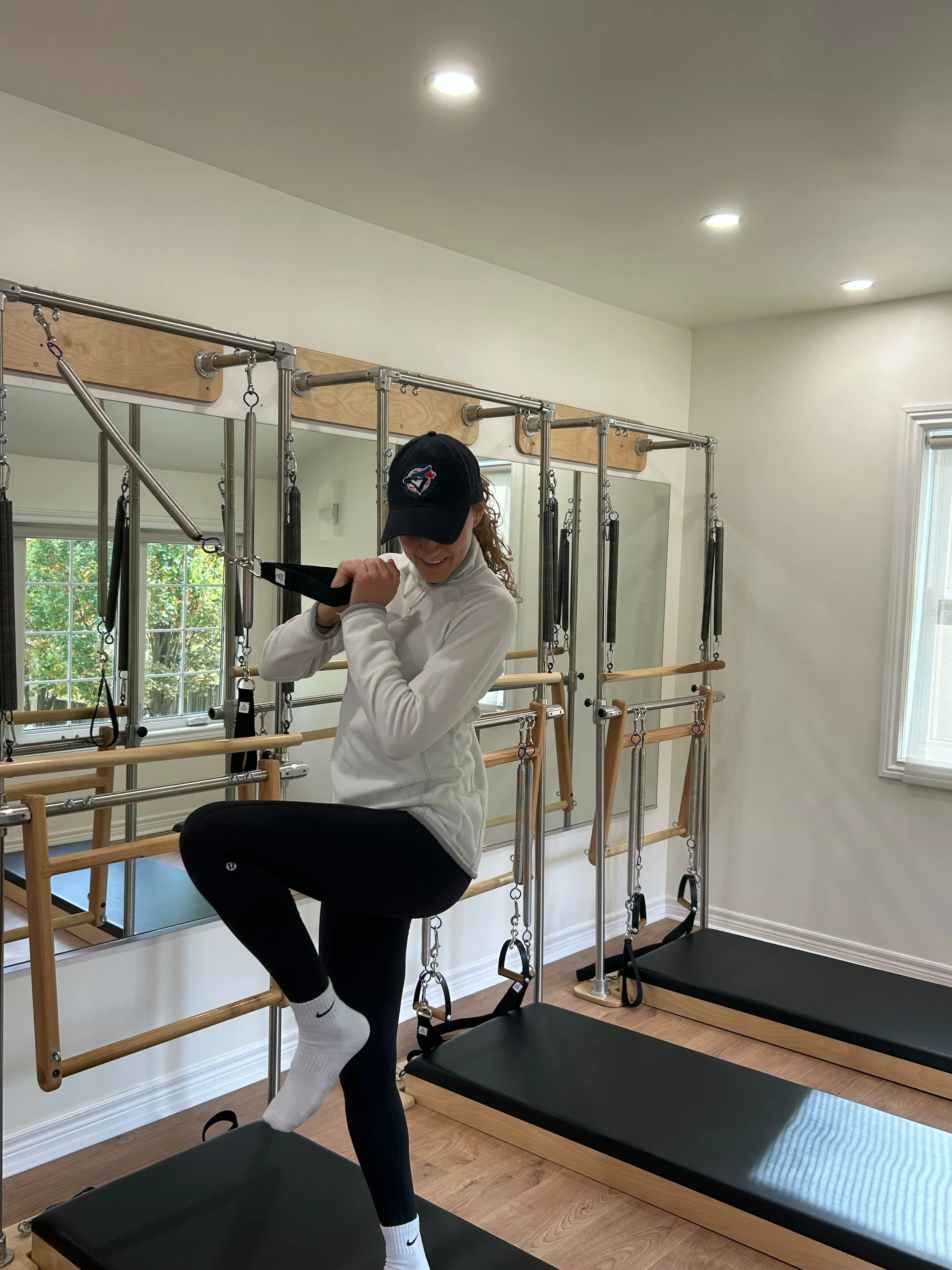 Woman using Pilates equipment in a home gym, wearing a black Nike cap and white jacket.