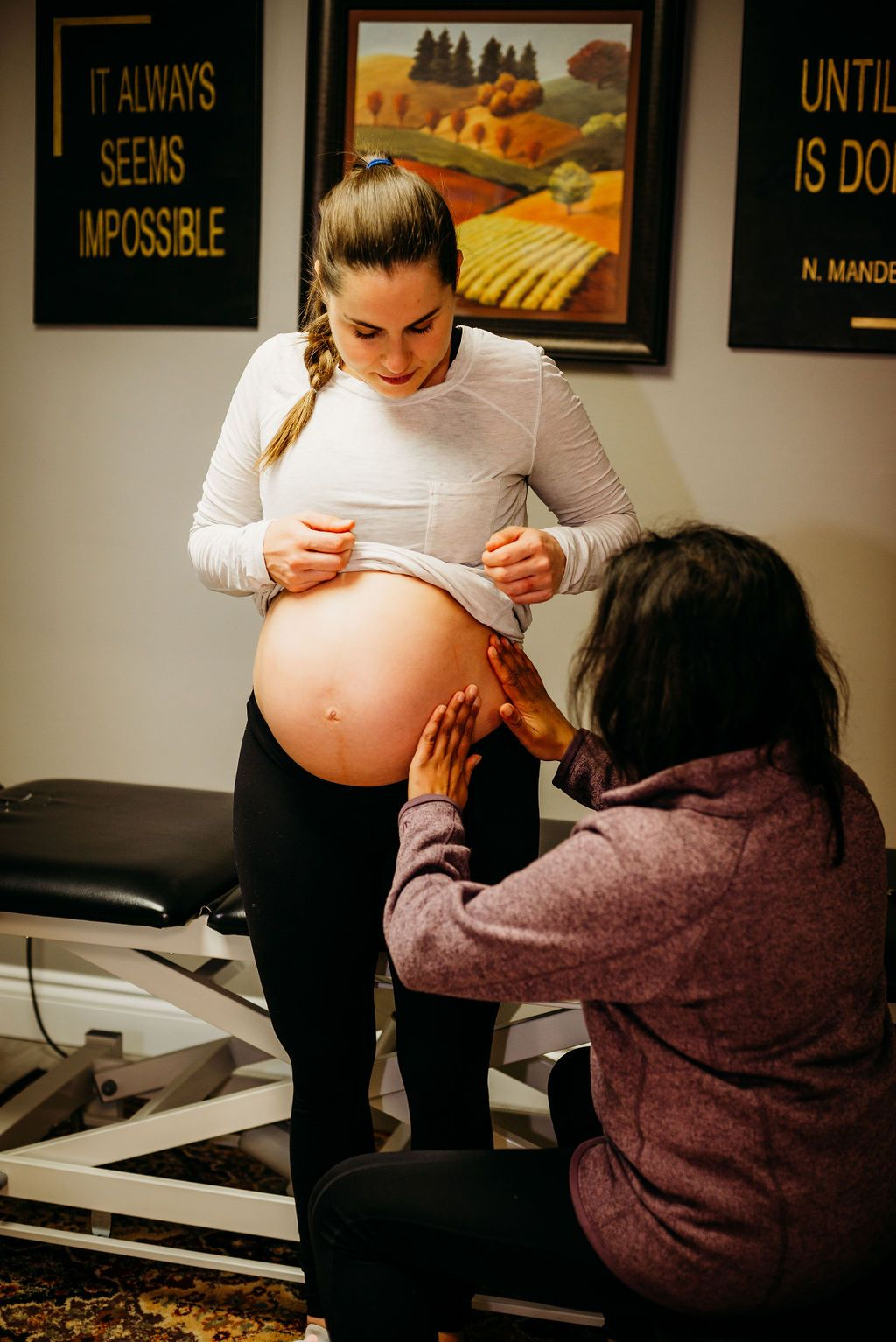 A pregnant woman in a white long-sleeve shirt lifts her shirt to reveal her belly while a woman kneeling in front of her touches her stomach, inside a room with framed artwork and motivational quotes on the wall.