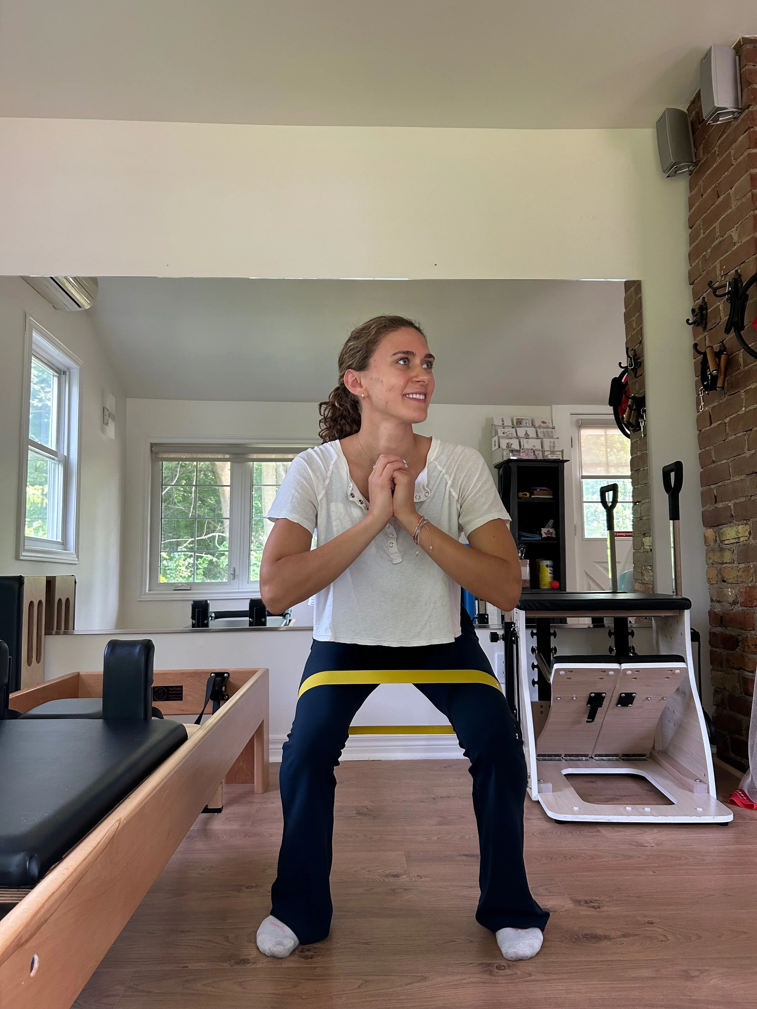A woman standing in a room with her legs apart and hands clasped in front of her chest, wearing striped socks and sweatpants that are bunched up at the ankles. The room has a large window, brick wall, and exercise equipment.