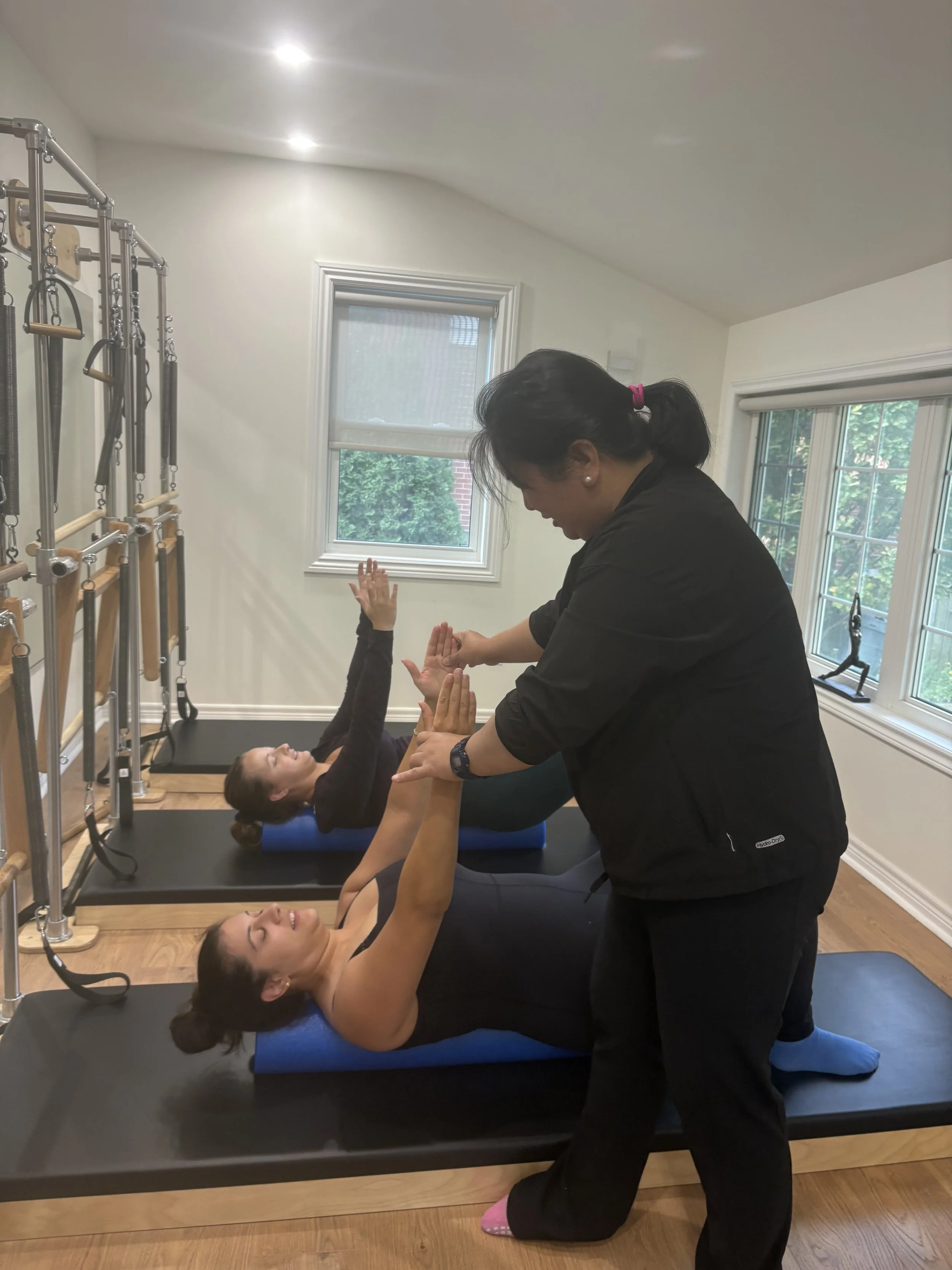 A woman lying on a Pilates reformer machine, smiling, while another woman assisting her prepares to perform an exercise, all in a bright room with windows and exercise equipment.