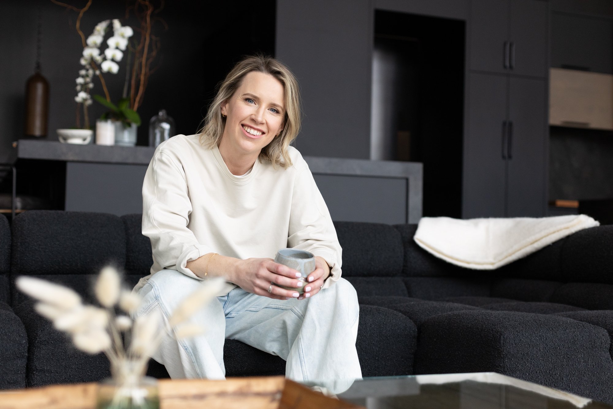 A smiling woman with blonde hair sitting on a black sofa, holding a ceramic mug, in a modern living room with dark walls and minimalist decor.