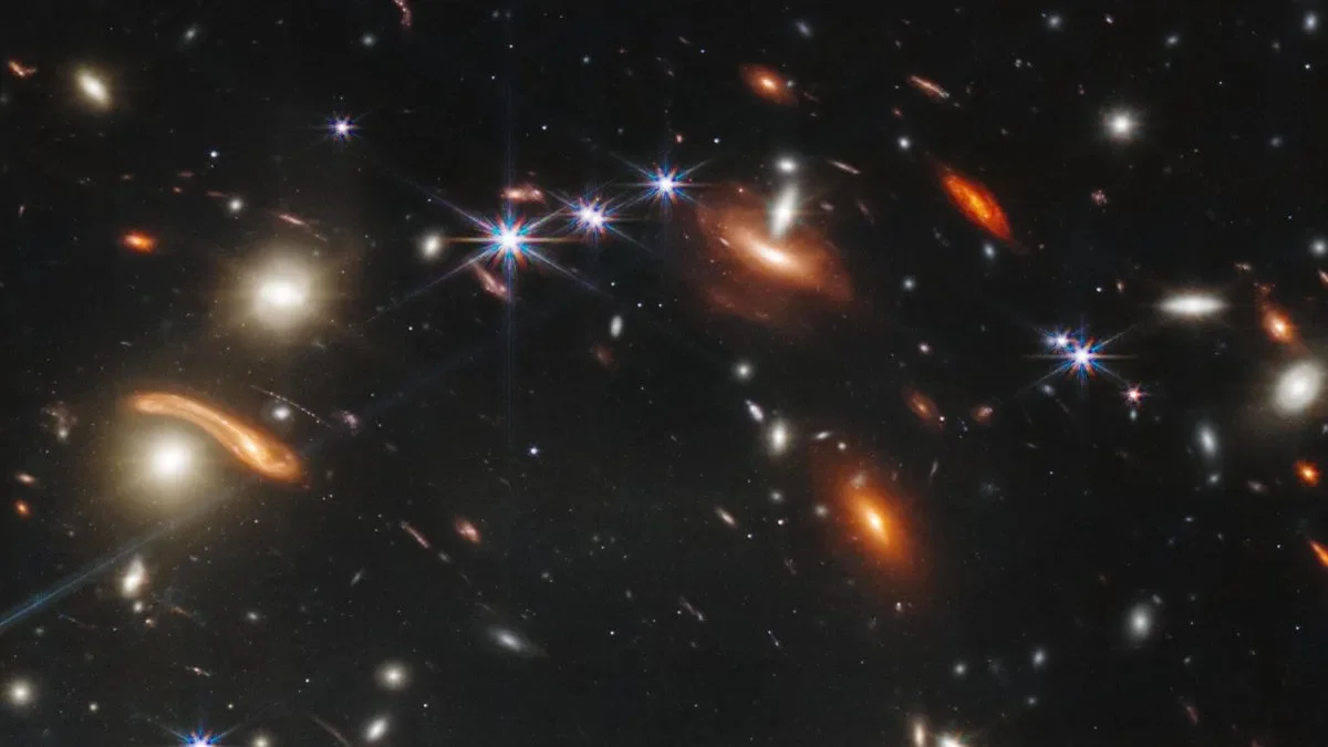 Deep space image showing numerous galaxies, some with bright, glowing centers and others with elongated or irregular shapes, set against a dark background with many faint stars.