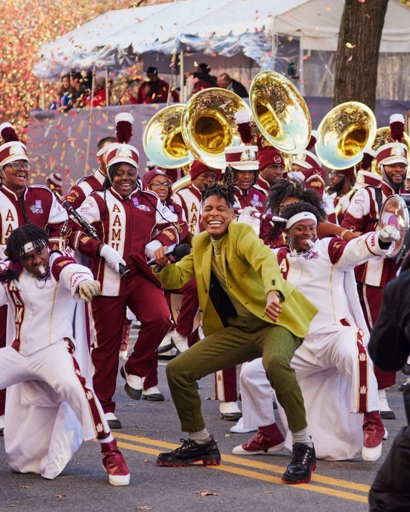 Marching band in maroon and white uniforms performing with sousaphones during a parade, with Jon Batiste in a green suit dancing in front.
