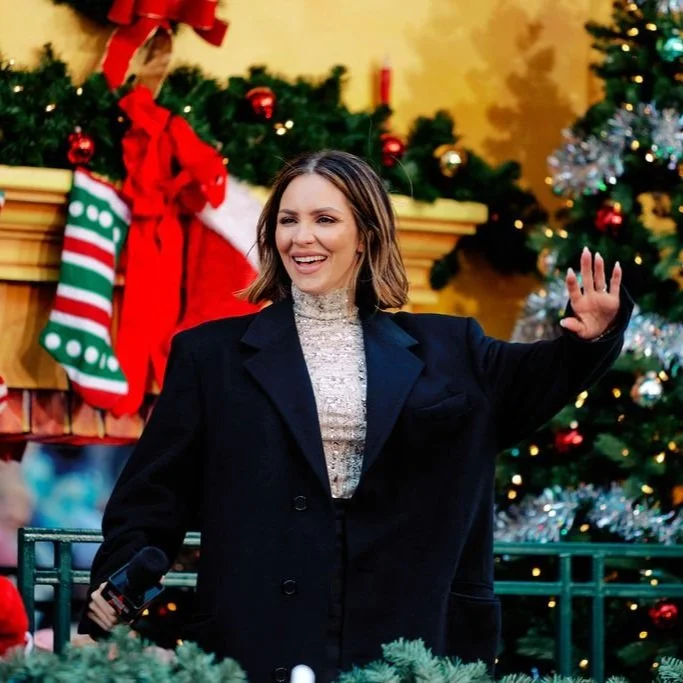 Katherine McPhee Foster wearing a black coat, holding a microphone, standing in front of a Christmas-themed background with garlands and stockings. The person is smiling and waving. Holiday decorations are visible.