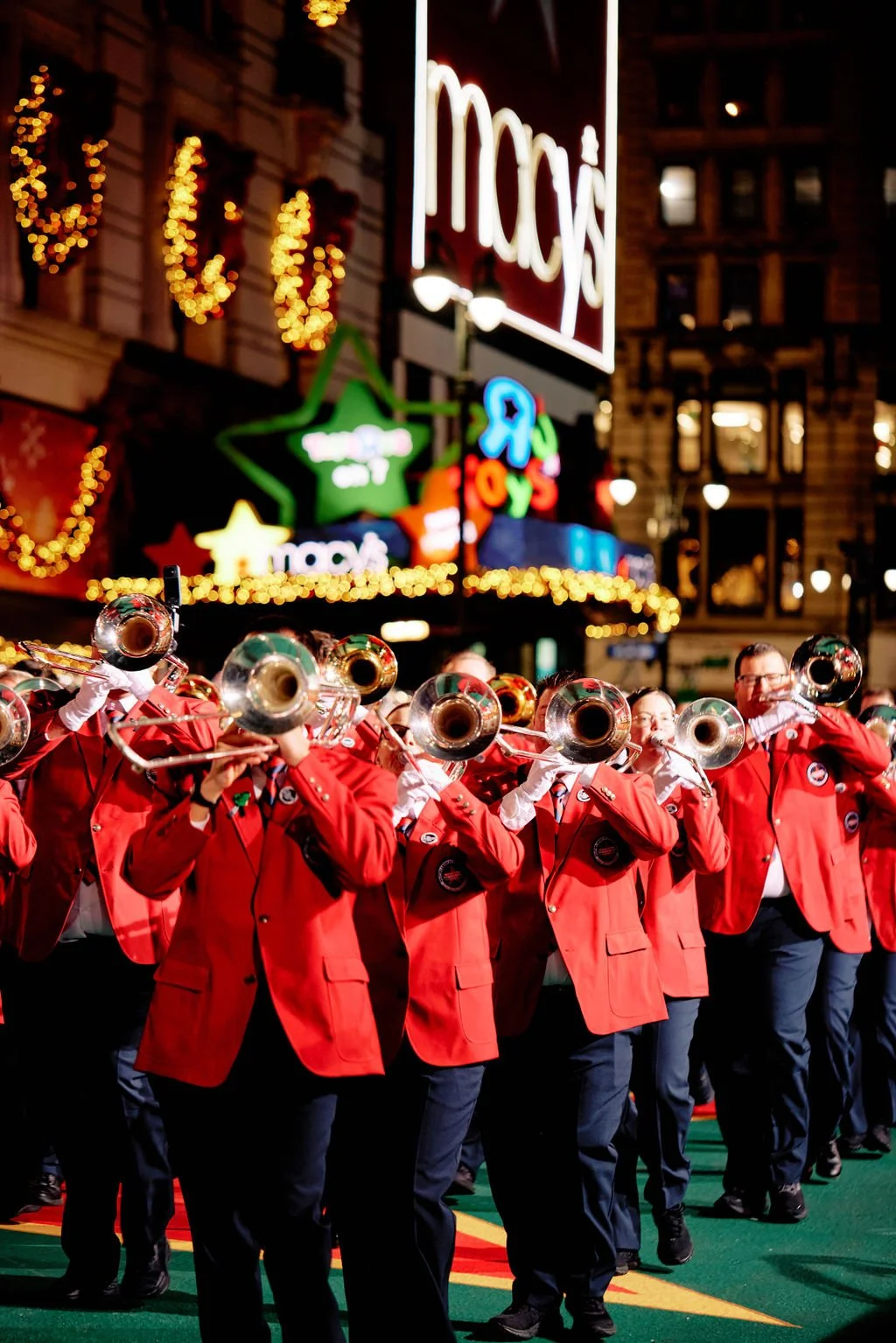 Marching band in red uniforms with trumpets in front of Macy's decorated with holiday lights.