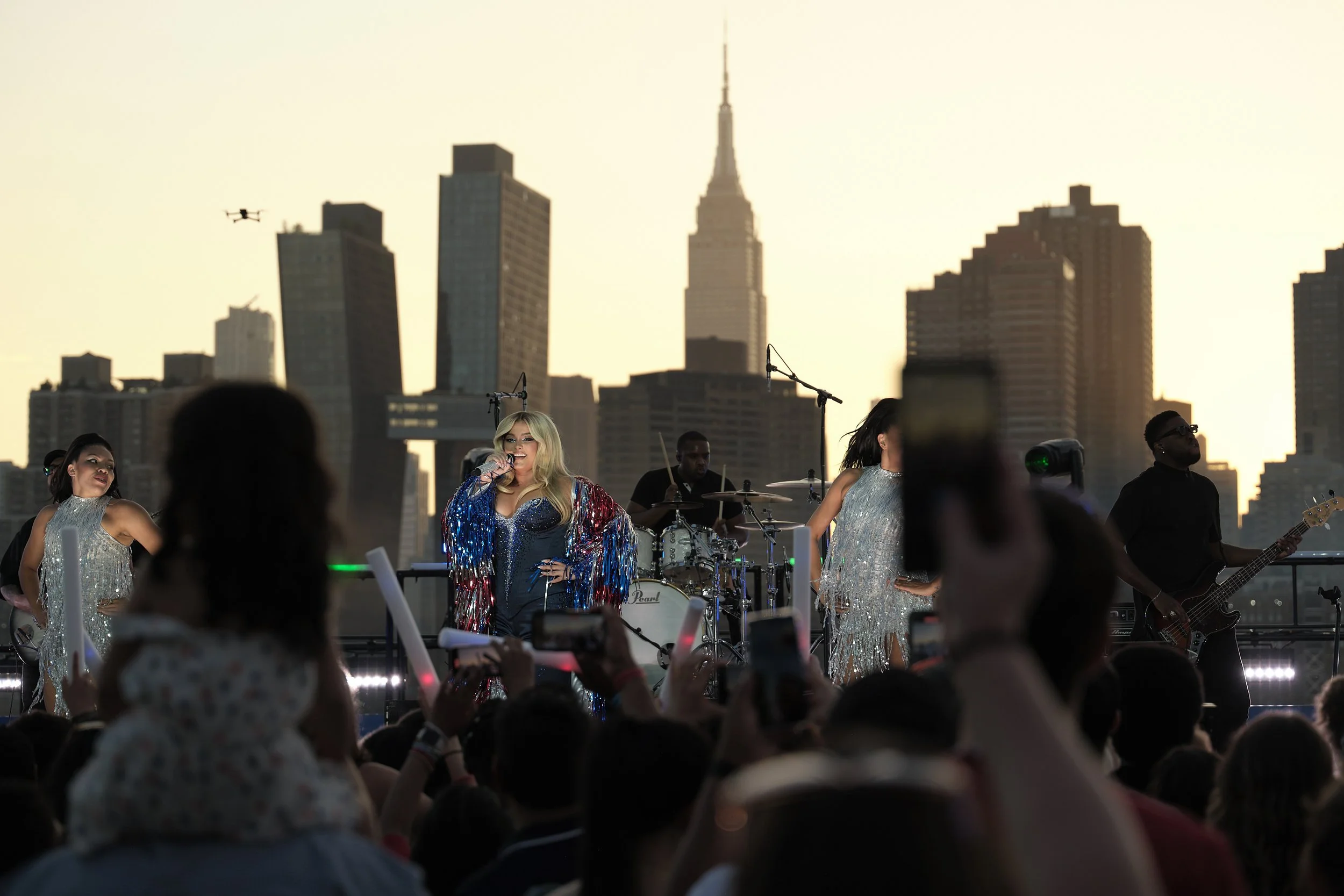 Outdoor concert with a singer performing on stage, surrounded by band members, in front of a city skyline at sunset.