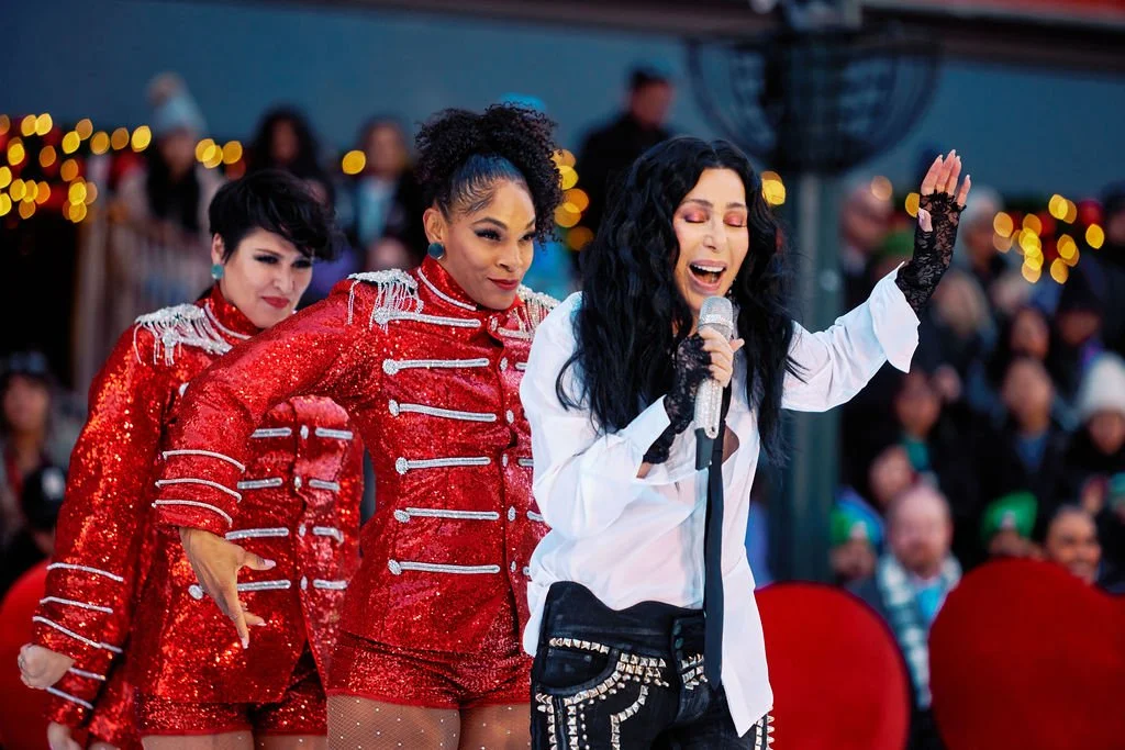 Cher performing on stage with two dancers in red sequin outfits, holding a microphone, with a festive background.