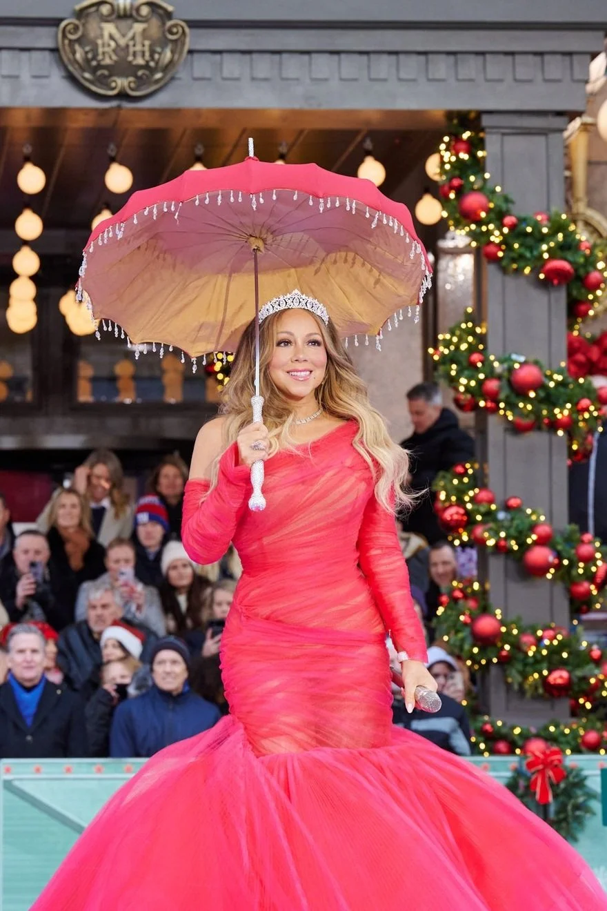 Mariah Carey in a red gown and tiara holds a pink umbrella. She is standing in front of Christmas decorations and a crowd of people.