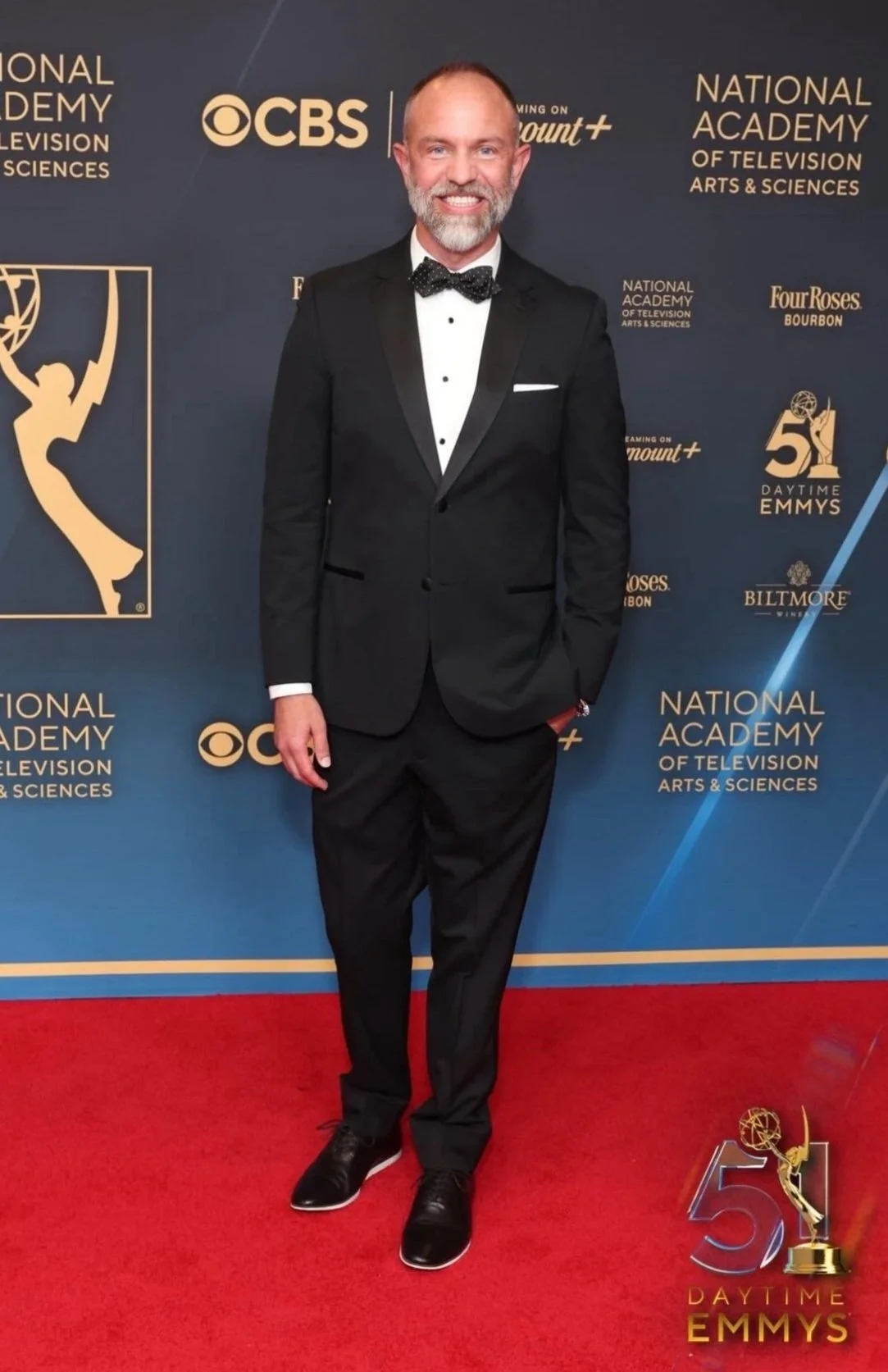 Man in a tuxedo at the 51st Daytime Emmys event, standing on a red carpet with Daytime Emmys signage in the background.