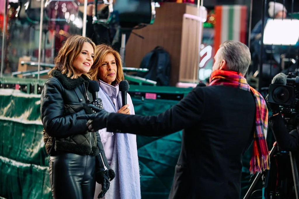 Hoda Kotb and Savannah Guthrie with microphones engaging in conversation on a set with a camera operator filming.