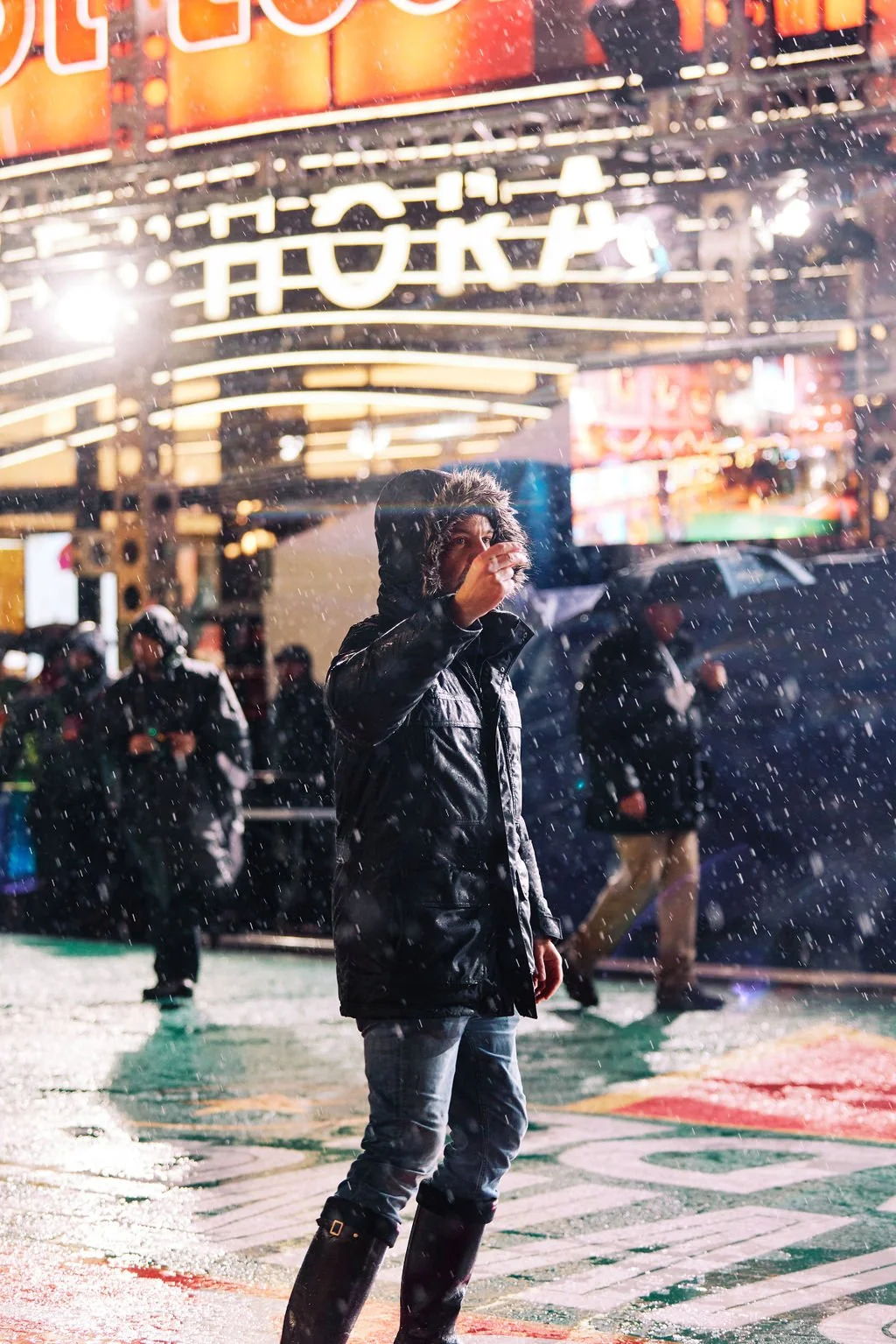 Person in a fur-lined hooded jacket standing in snowy urban environment with bright lights and signage in the background.