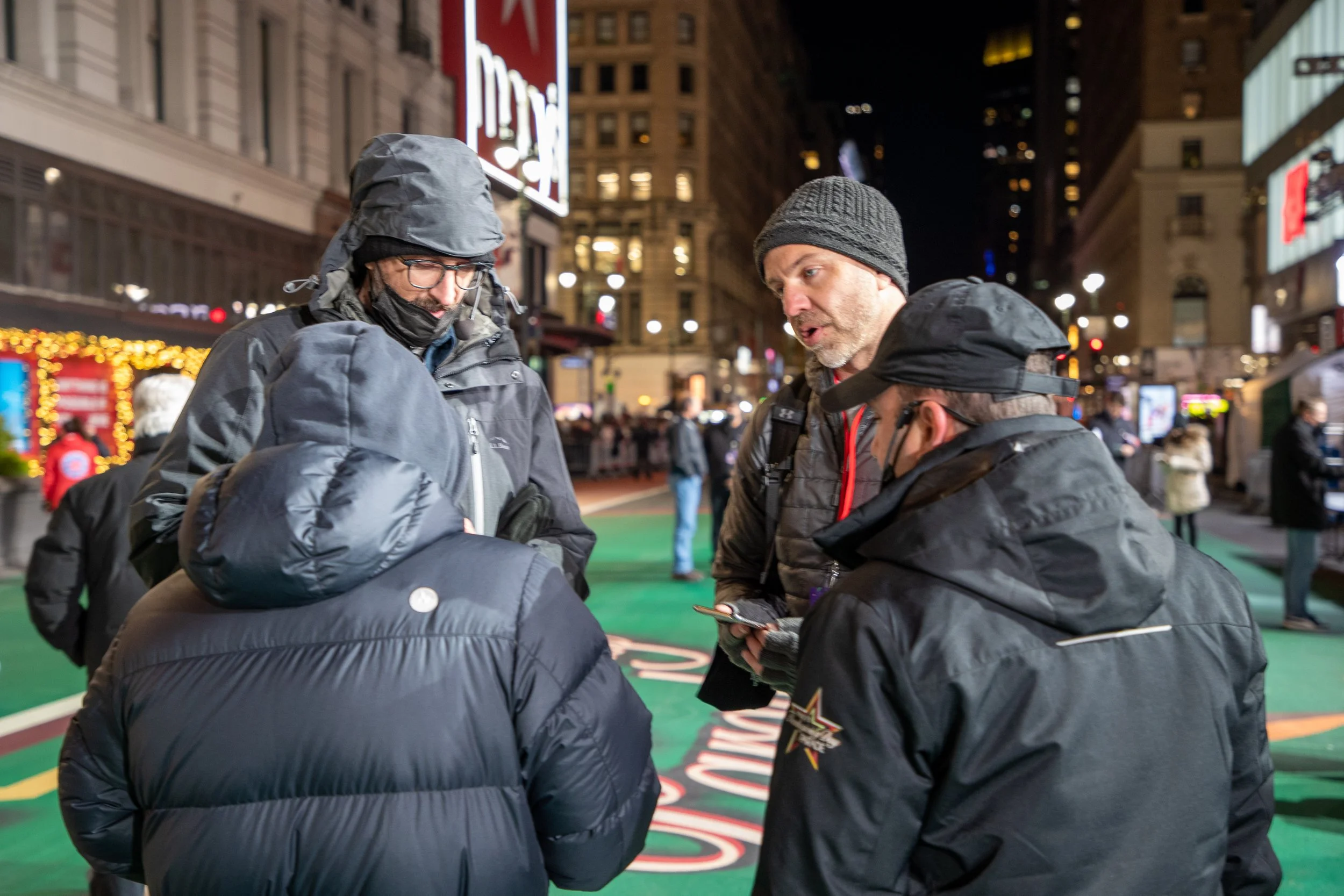 Group of four people wearing winter jackets and hats, standing on a city street at night near holiday decorations.