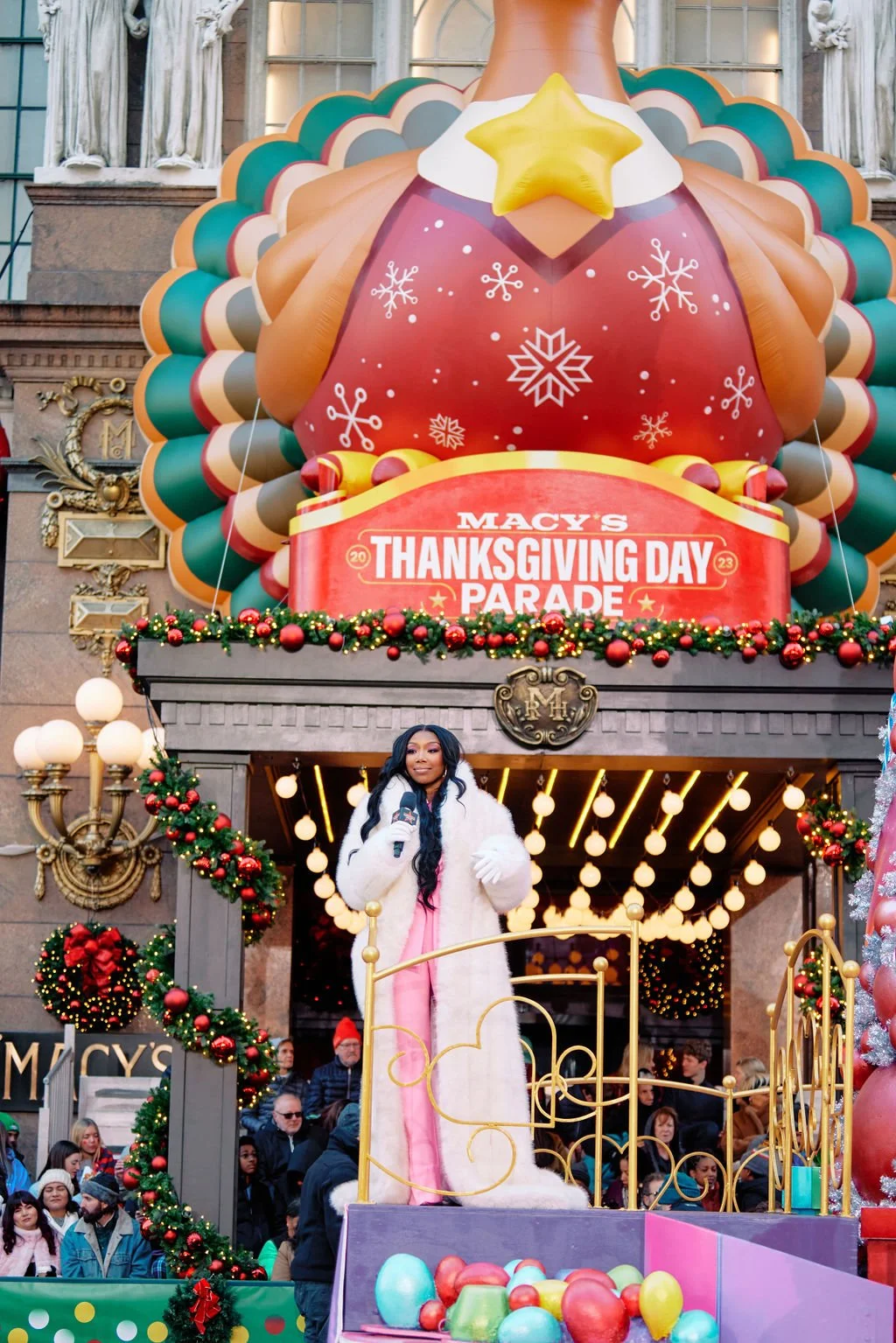 A person in a white coat stands on a float at Macy's Thanksgiving Day Parade in front of a large turkey balloon and festive decor.