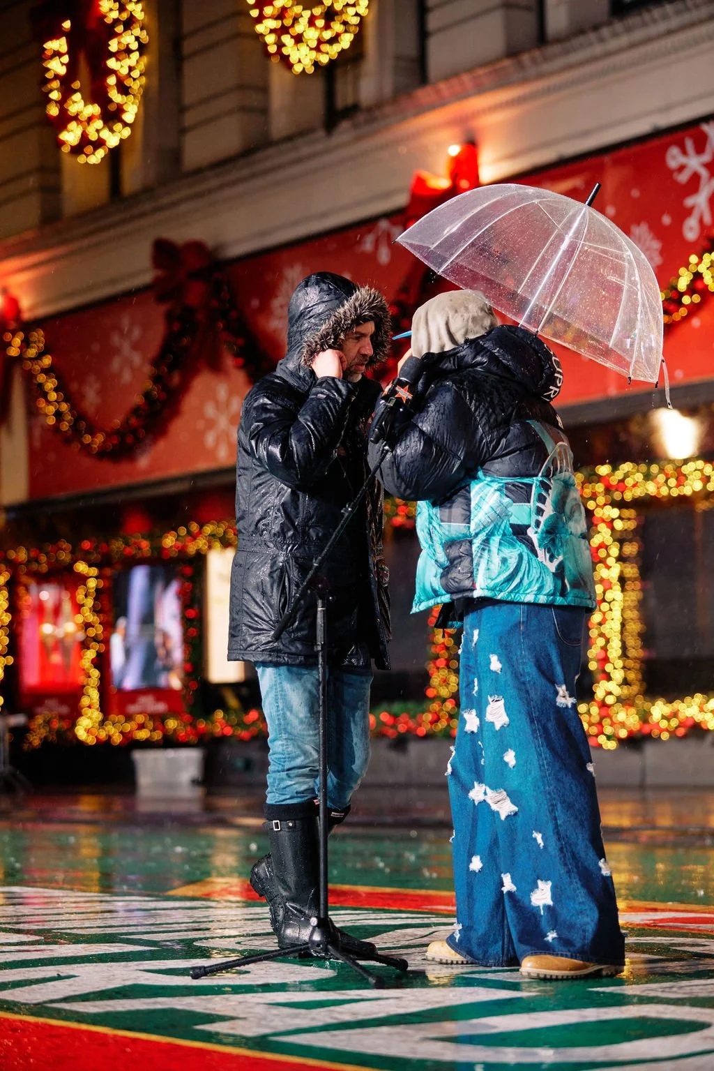 Two people standing close together on a decorated stage, one holding a transparent umbrella. They are wearing winter jackets with hoods and jeans. The background features festive lights and decor.