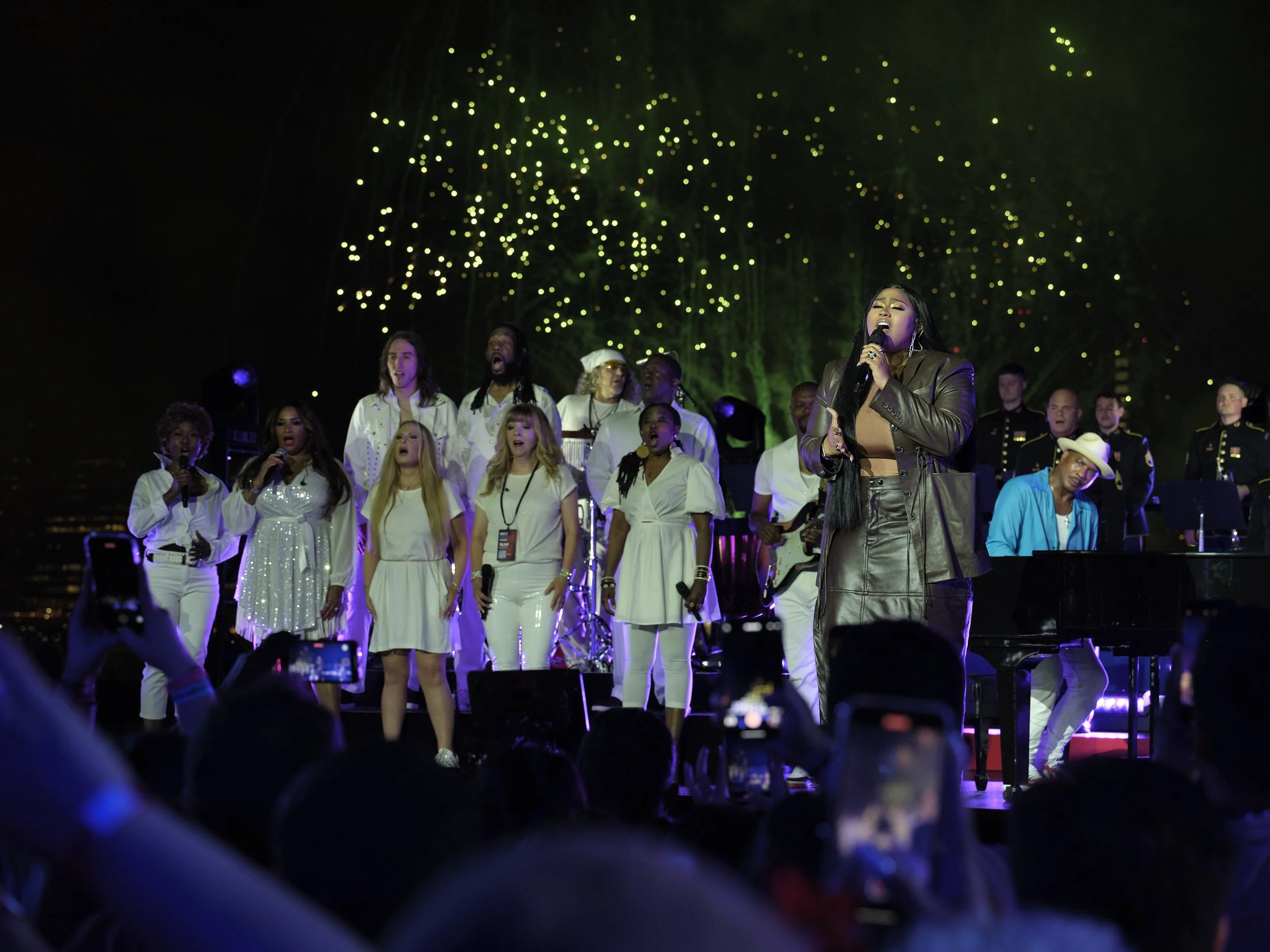 A group of performers on stage with fireworks in the background, featuring a singer in a brown leather outfit holding a microphone, surrounded by a choir dressed in white and a pianist wearing a blue jacket.