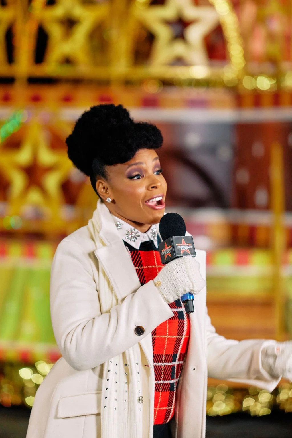 Person holding a microphone, wearing a red plaid sweater and white coat, with a festive background.