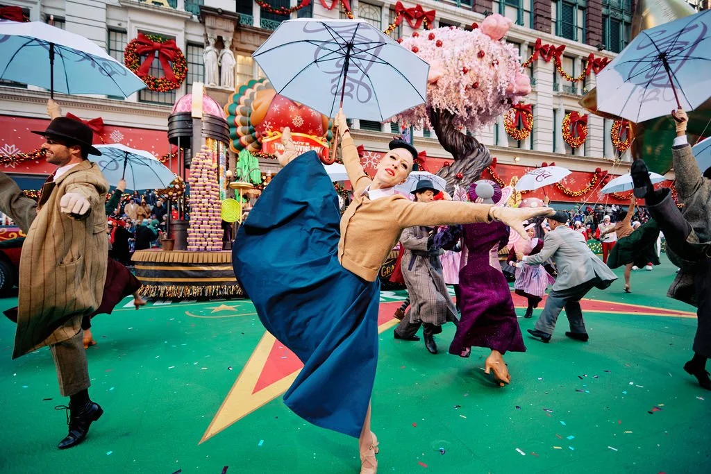 Wonka Movie Performers in colorful costumes dance with umbrellas at a festive event, possibly a parade. The background features a large, decorated float and holiday decorations, including wreaths and bows.