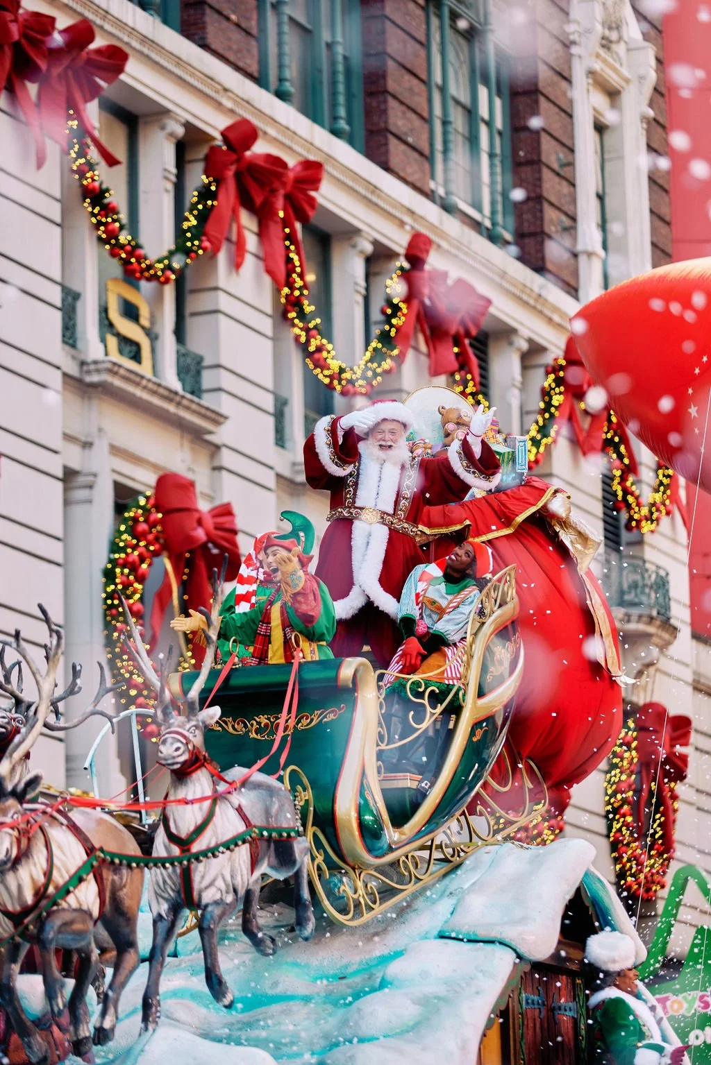 Santa Claus waving from a festive sleigh with reindeer during a Christmas parade, surrounded by elves and holiday decorations, with snow falling and a decorated building in the background.