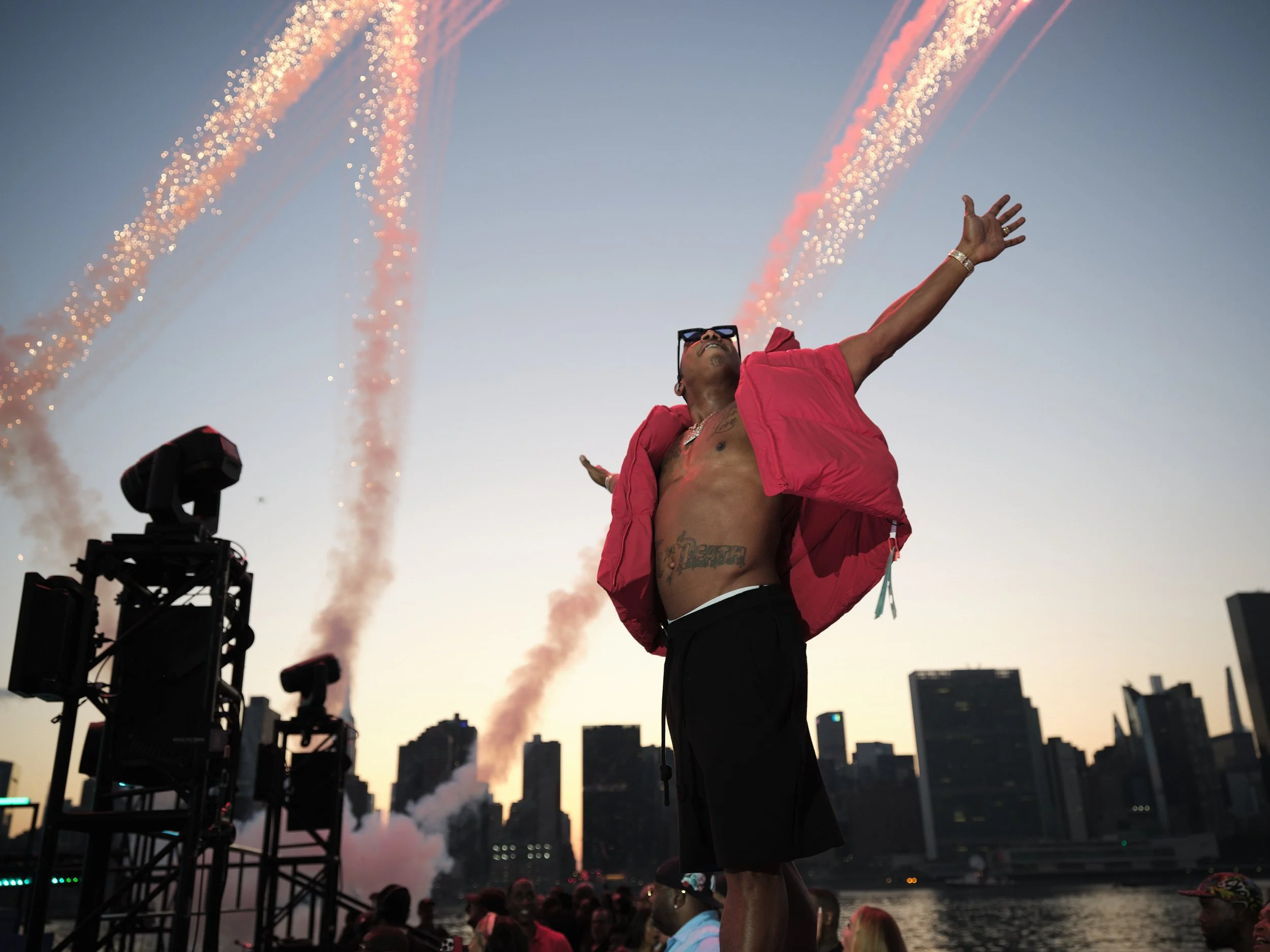 Nelly celebrating with arms raised, fireworks in background, city skyline at dusk.