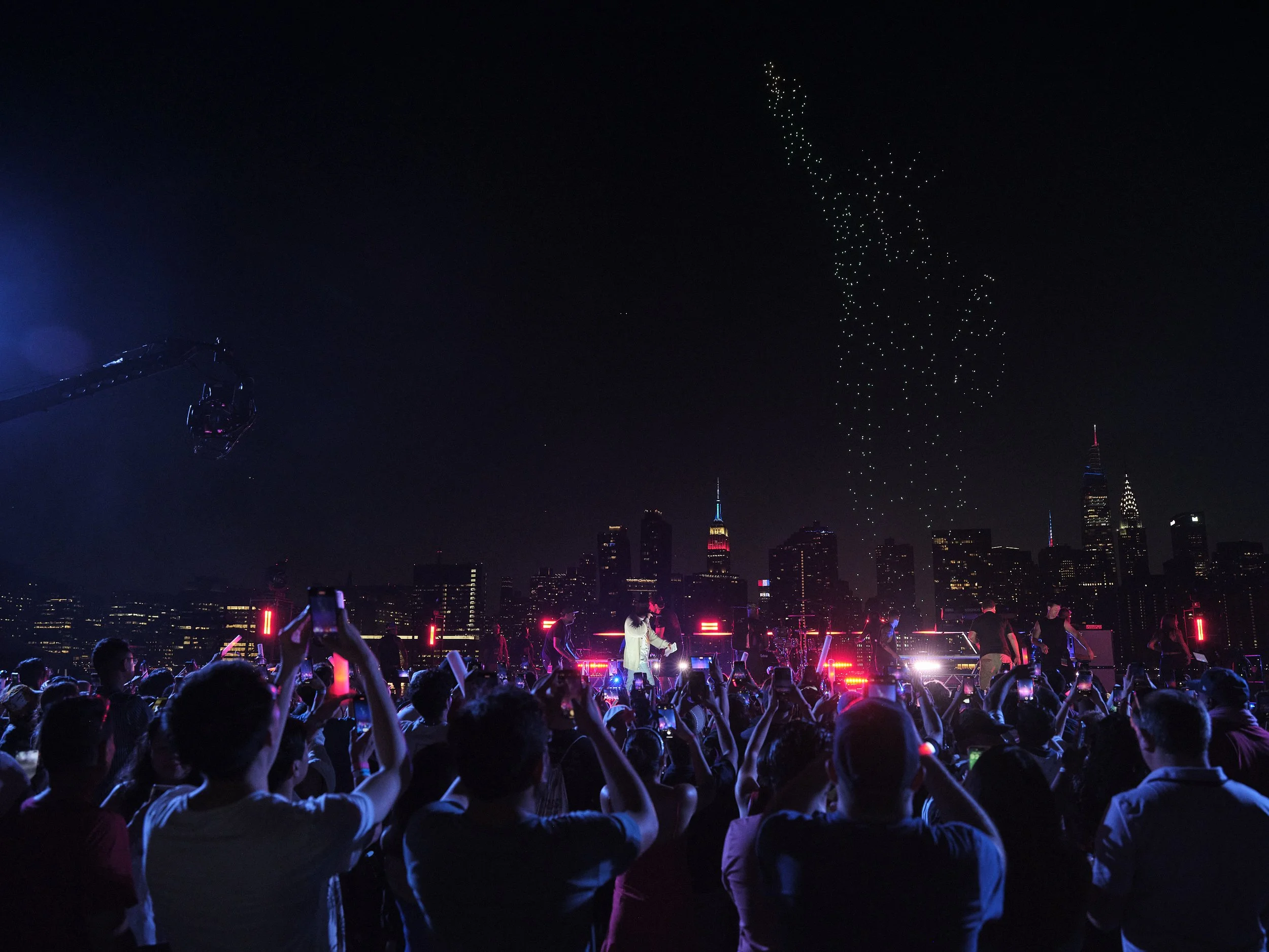 Concert crowd with city skyline and night sky featuring drone light display forming Statue of Liberty silhouette.