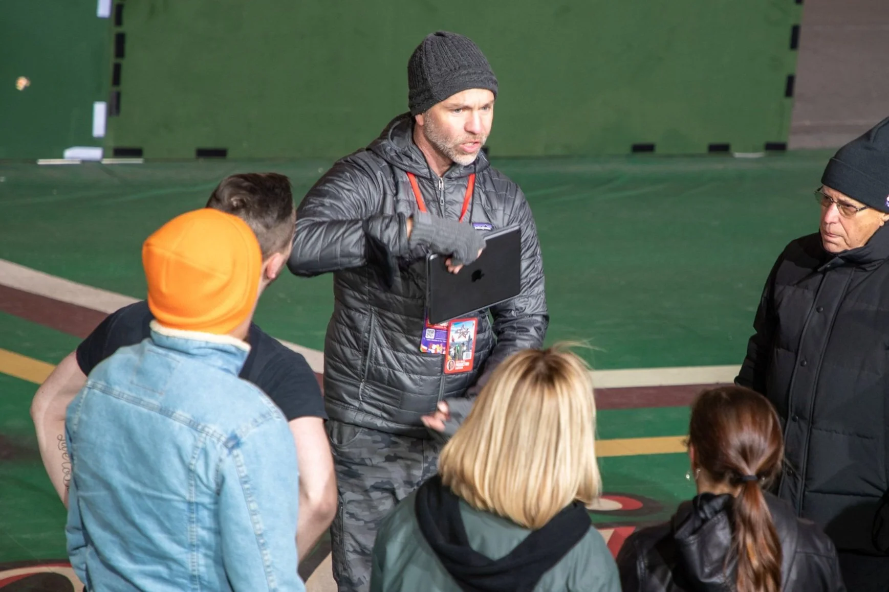 A group of people stand indoors, one man in a gray jacket and beanie holding a tablet and speaking to the others. One person wears an orange beanie, while others wear dark winter clothing. They appear to be on a sports field or stadium setting.