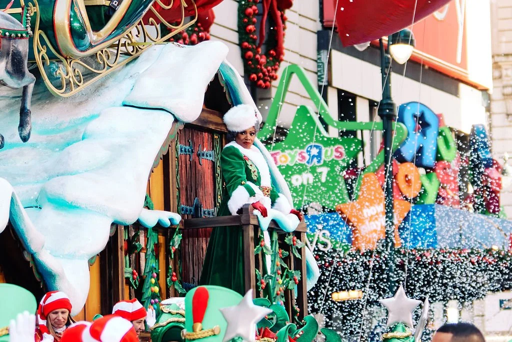 Christmas parade float featuring a Sheryl Lee Ralph in green and white attire, surrounded by festive decorations, with snow-like confetti falling.