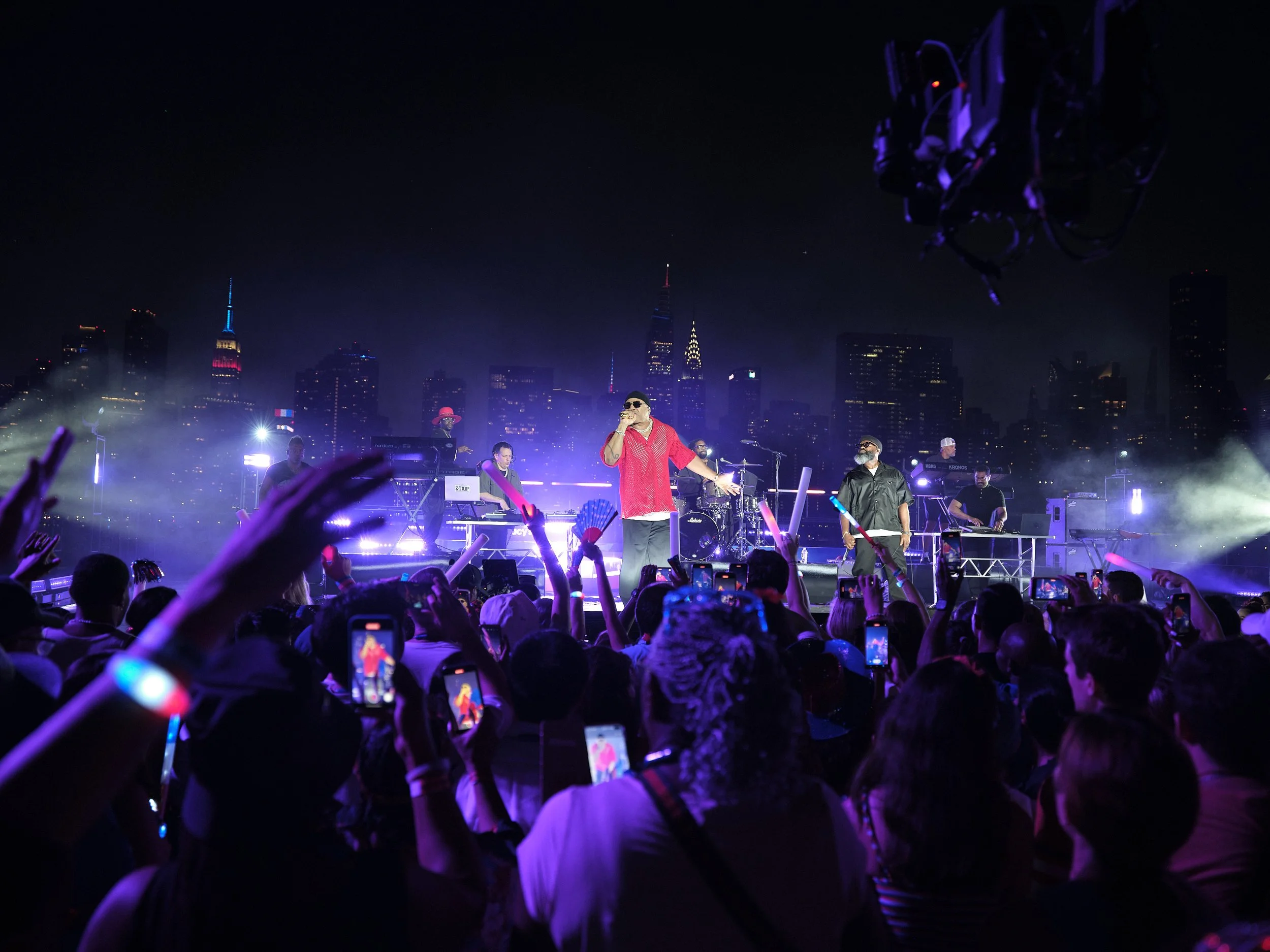 Nighttime outdoor concert with a performer on stage under colorful lights, surrounded by a crowd with raised hands and smartphones, against a city skyline backdrop.