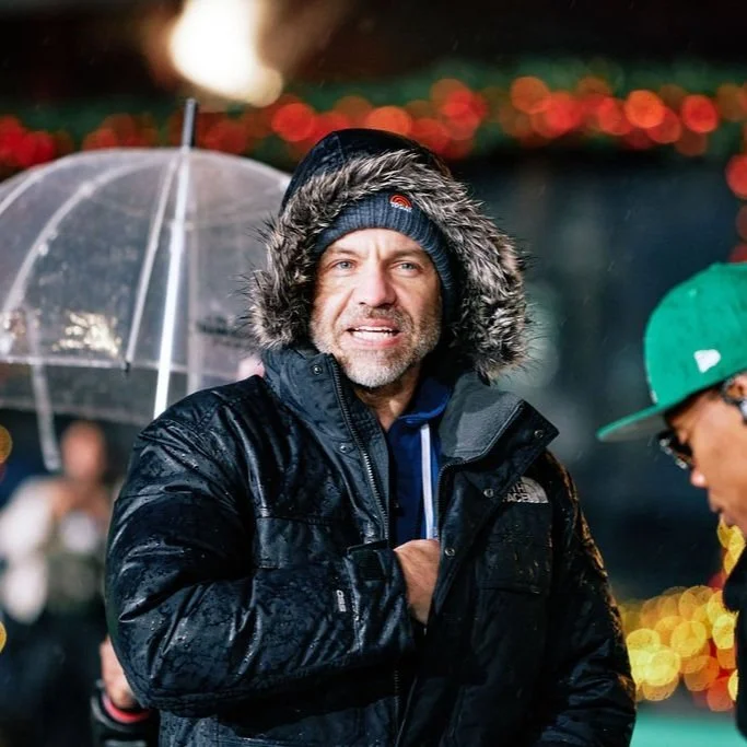 Man wearing a hooded winter jacket and knit cap, standing outdoors in rainy weather, holding an umbrella. Festive lights in the background.