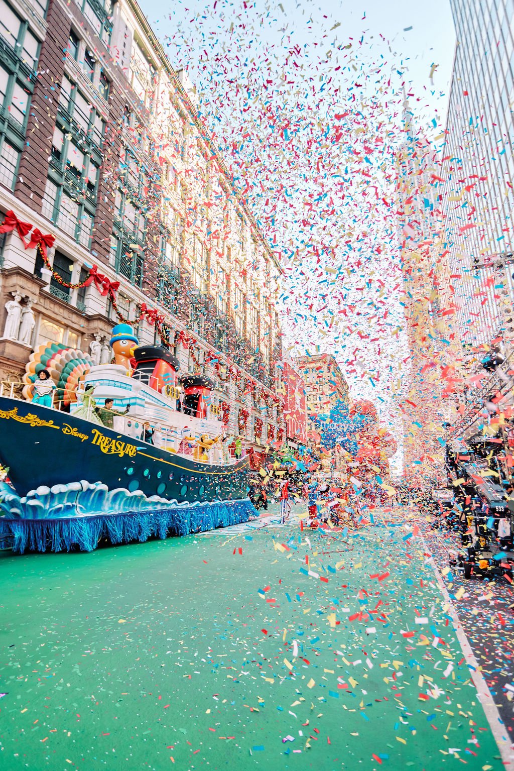 A festive parade scene on a city street with colorful confetti falling, a decorated float resembling a ship labeled "Disney Treasure," and surrounding buildings adorned with holiday decorations.