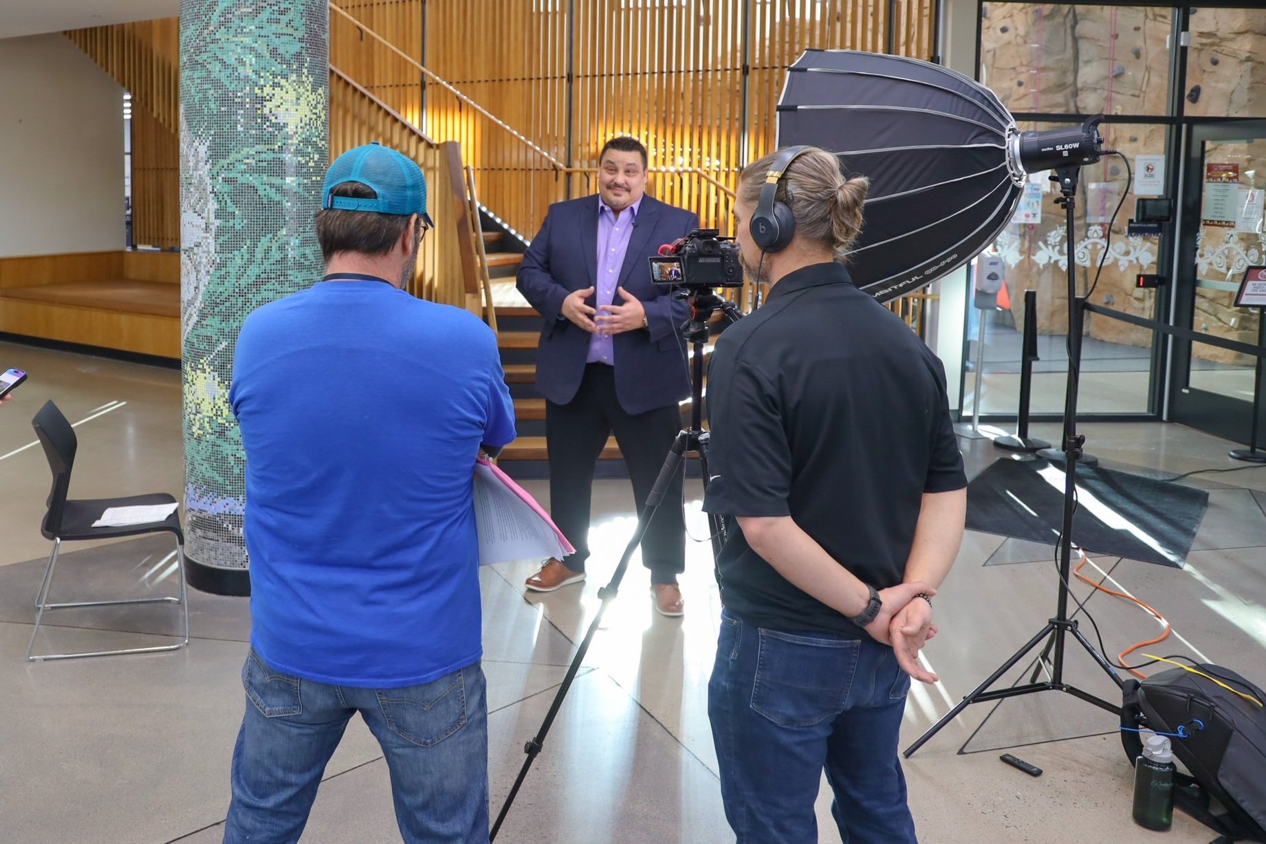 A man in a suit giving an interview to a camera crew inside a modern building with wooden accents and glass walls.