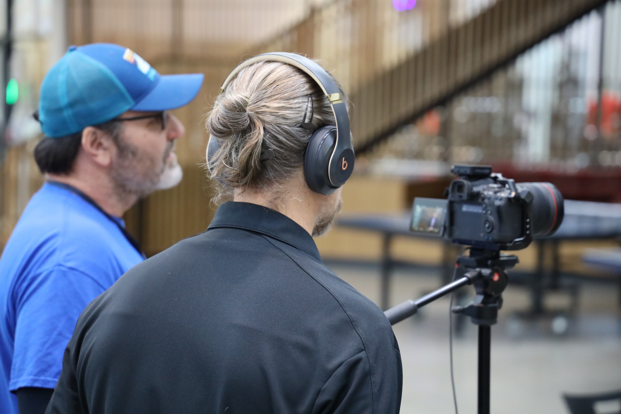 Two men standing backstage, one wearing a blue cap and shirt, the other wearing headphones and a black shirt, operating a camera on a tripod.