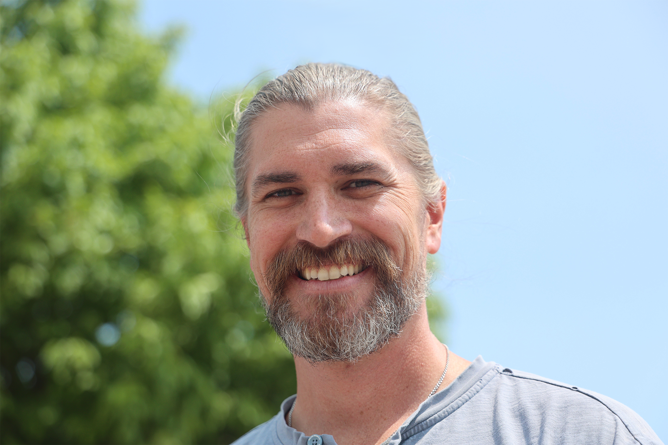 Close-up of a smiling man with gray hair and beard, wearing a gray shirt, outdoors with green trees and blue sky in the background.