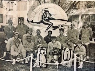 Group photo of U.S. Navy personnel in front of a mural featuring a sailor jumping out of the water with a rope, with a building and trees in the background.