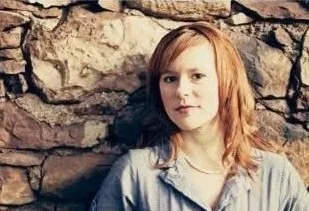 A young girl with red hair standing against a stone wall, wearing a gray shirt.