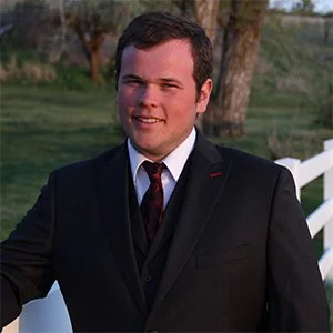 A young man in a black suit and tie smiling outdoors with trees and a white fence behind him.