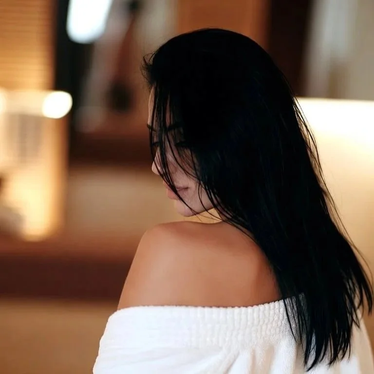 A woman with long black hair looking down, wearing a white off-the-shoulder top, in a softly lit indoor setting.
