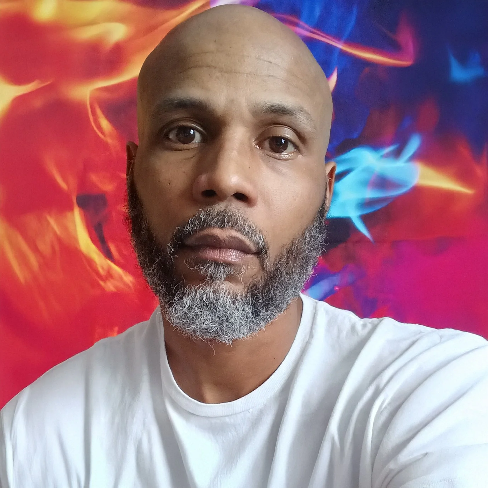 Close-up of a man with a beard and mustache wearing a white t-shirt, standing in front of a colorful abstract background with red, orange, blue, and purple swirling patterns.