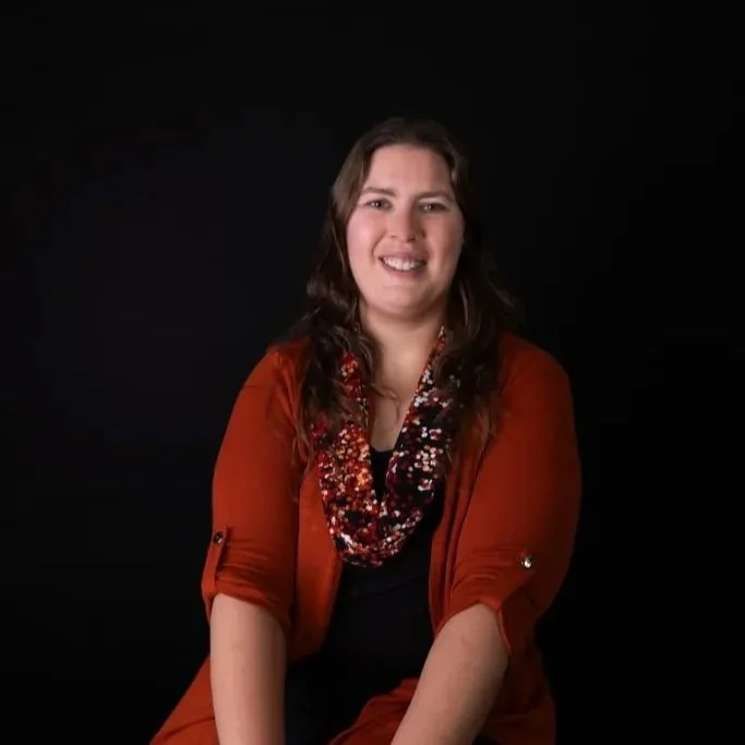A woman with long brown hair smiling, wearing a rust-colored jacket and a multi-colored, sequined scarf against a black background.