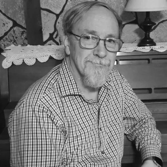 A middle-aged man with glasses, a beard, and light hair, wearing a checkered shirt, sitting indoors near a wooden piano with a lace doily and a lamp in the background.