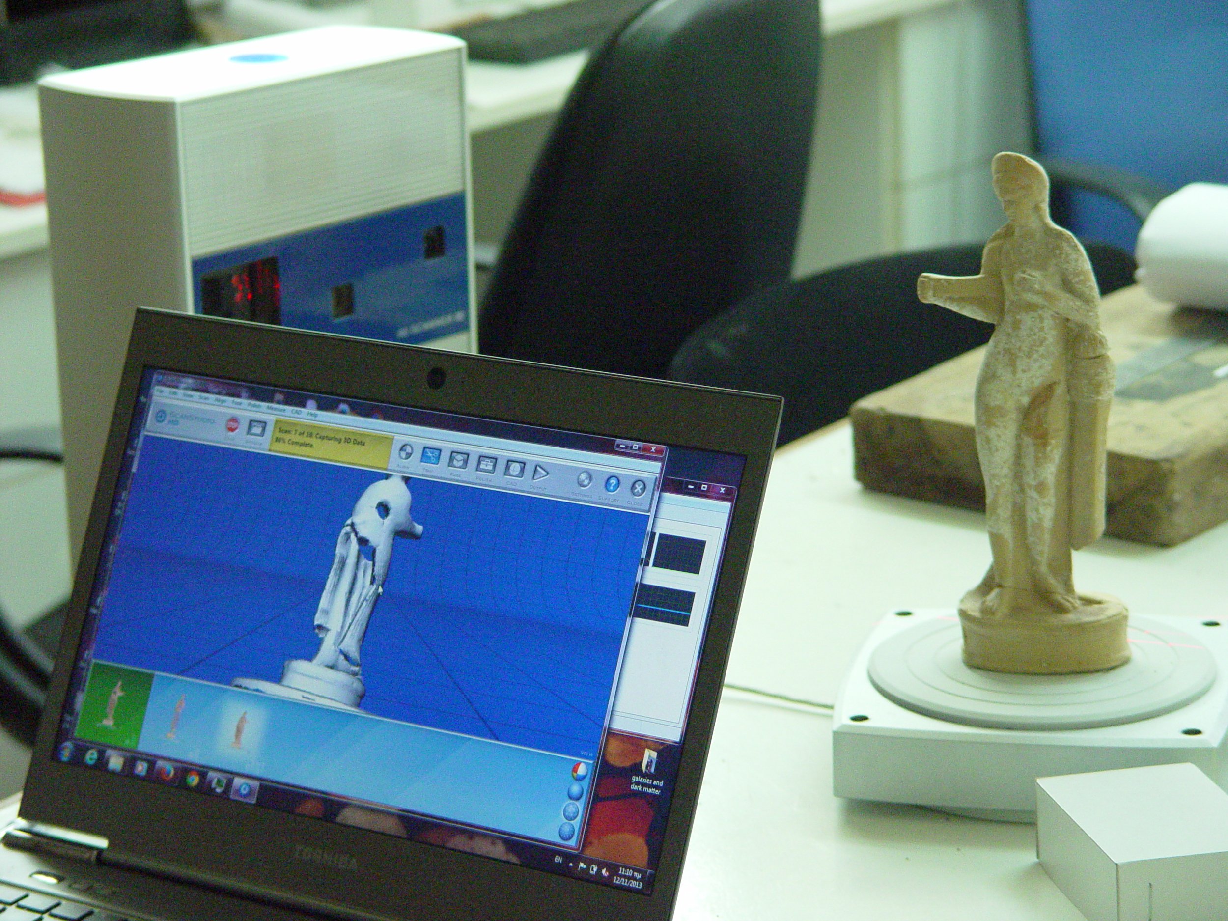 A computer screen displaying a 3D scan of a classical sculpture of a woman, with a physical clay sculpture of the same woman on a rotating platform beside it.