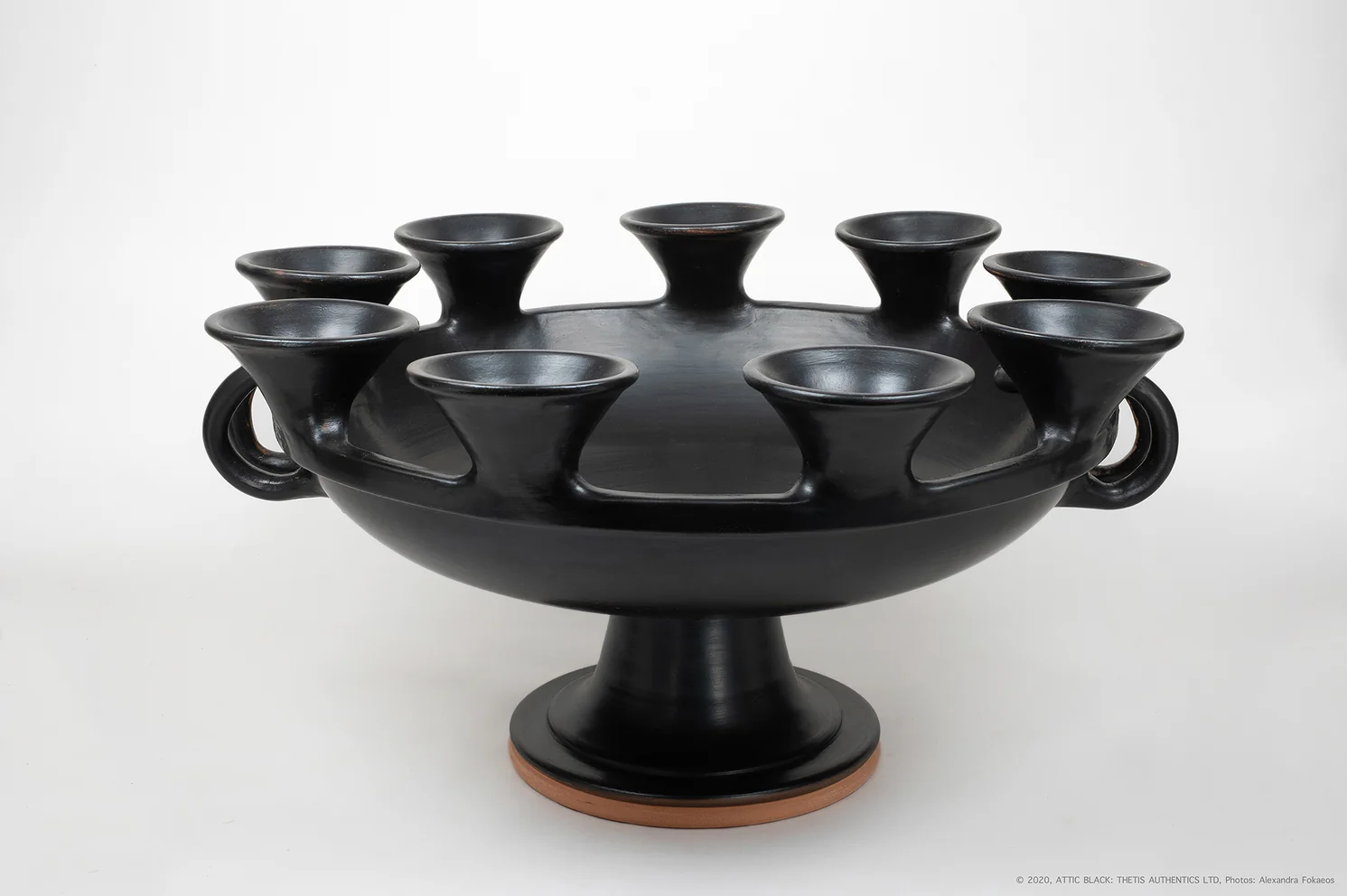 A black ceramic candle holder with ten small cups arranged around a central basin, mounted on a pedestal base.